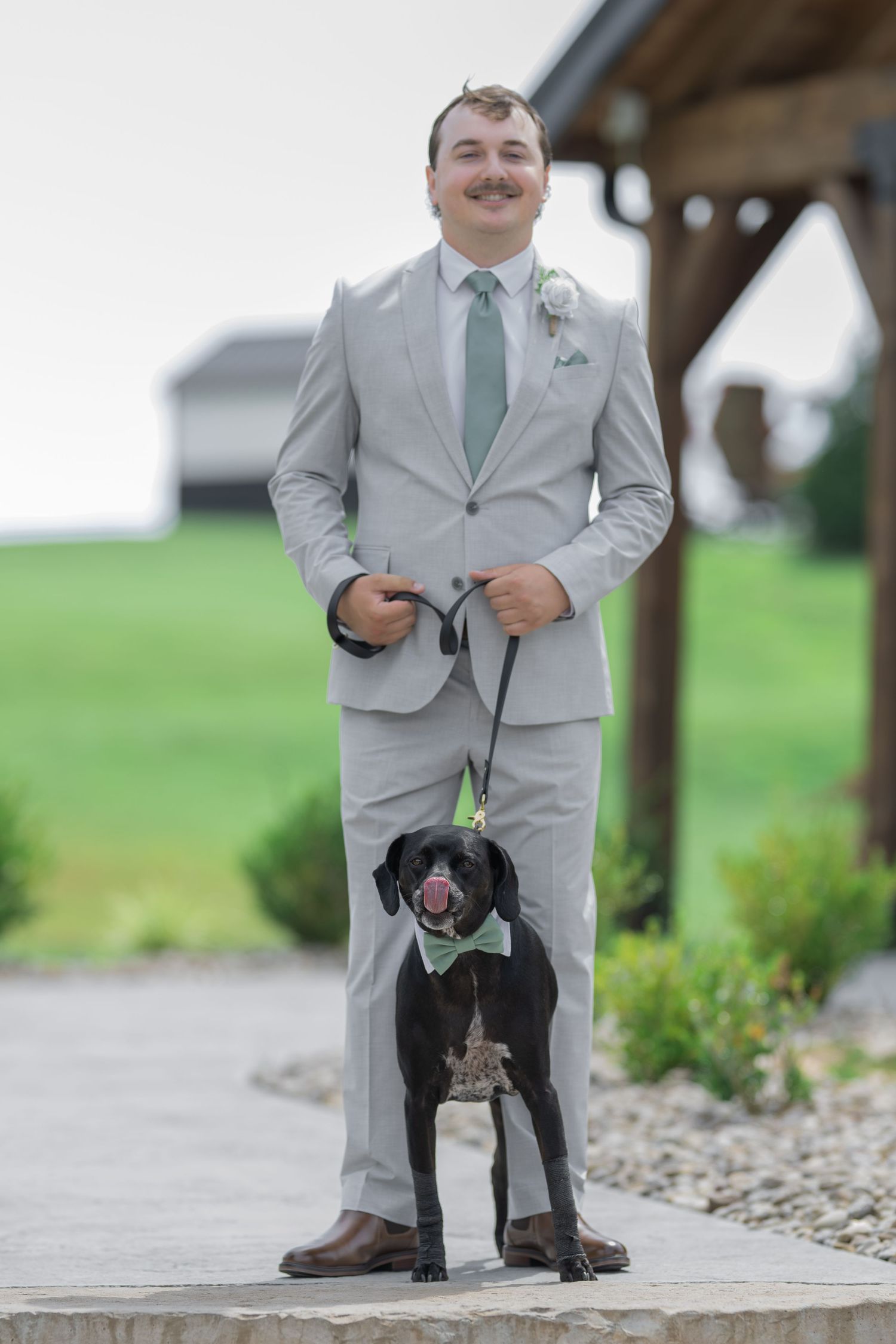 Smiling groom in gray suit with mint tie holds leash of black dog wearing matching bow tie at outdoor wedding venue.