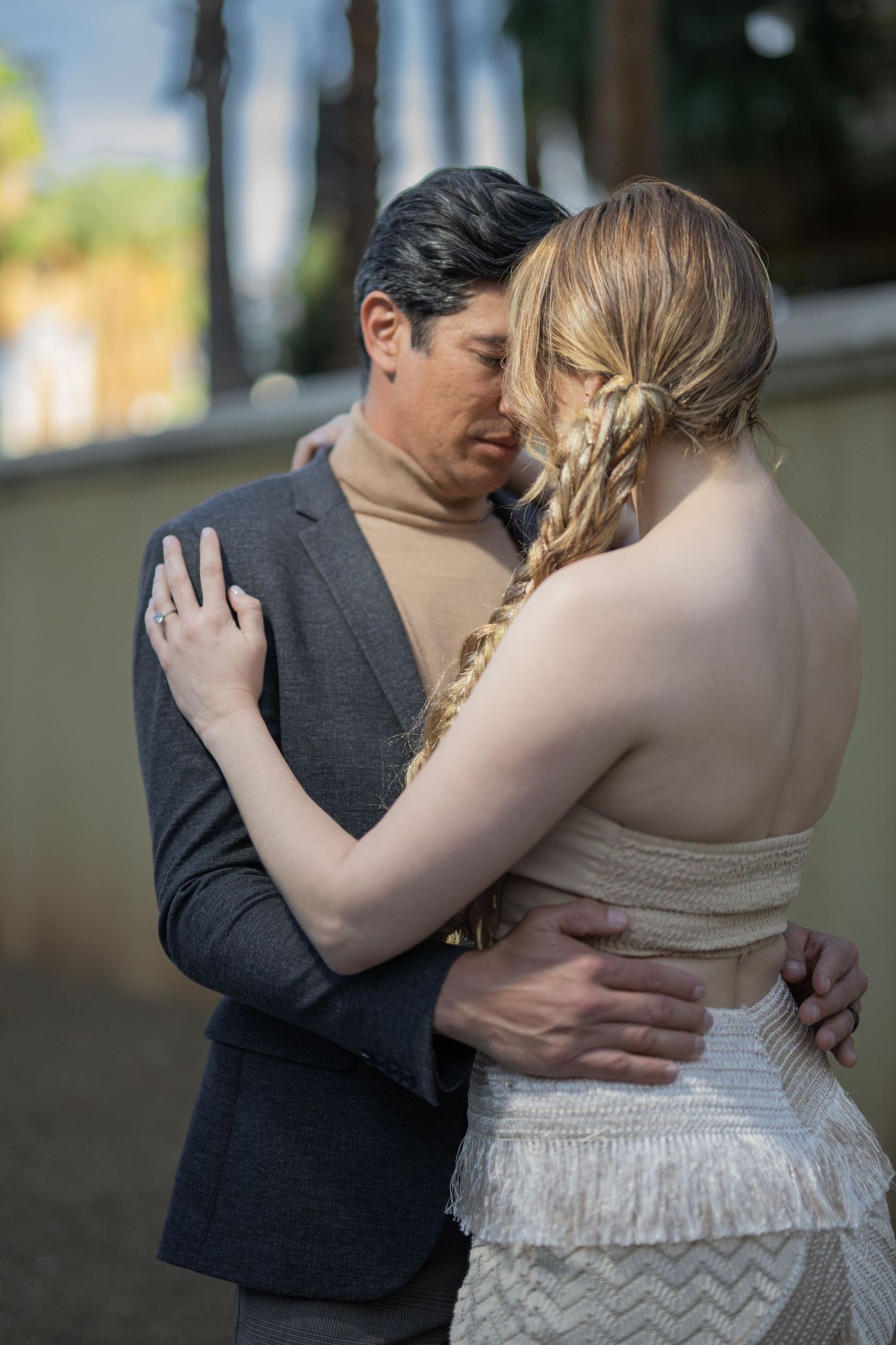 Couple sharing an intimate embrace, woman in white fringe dress with braided hair, man in gray blazer.