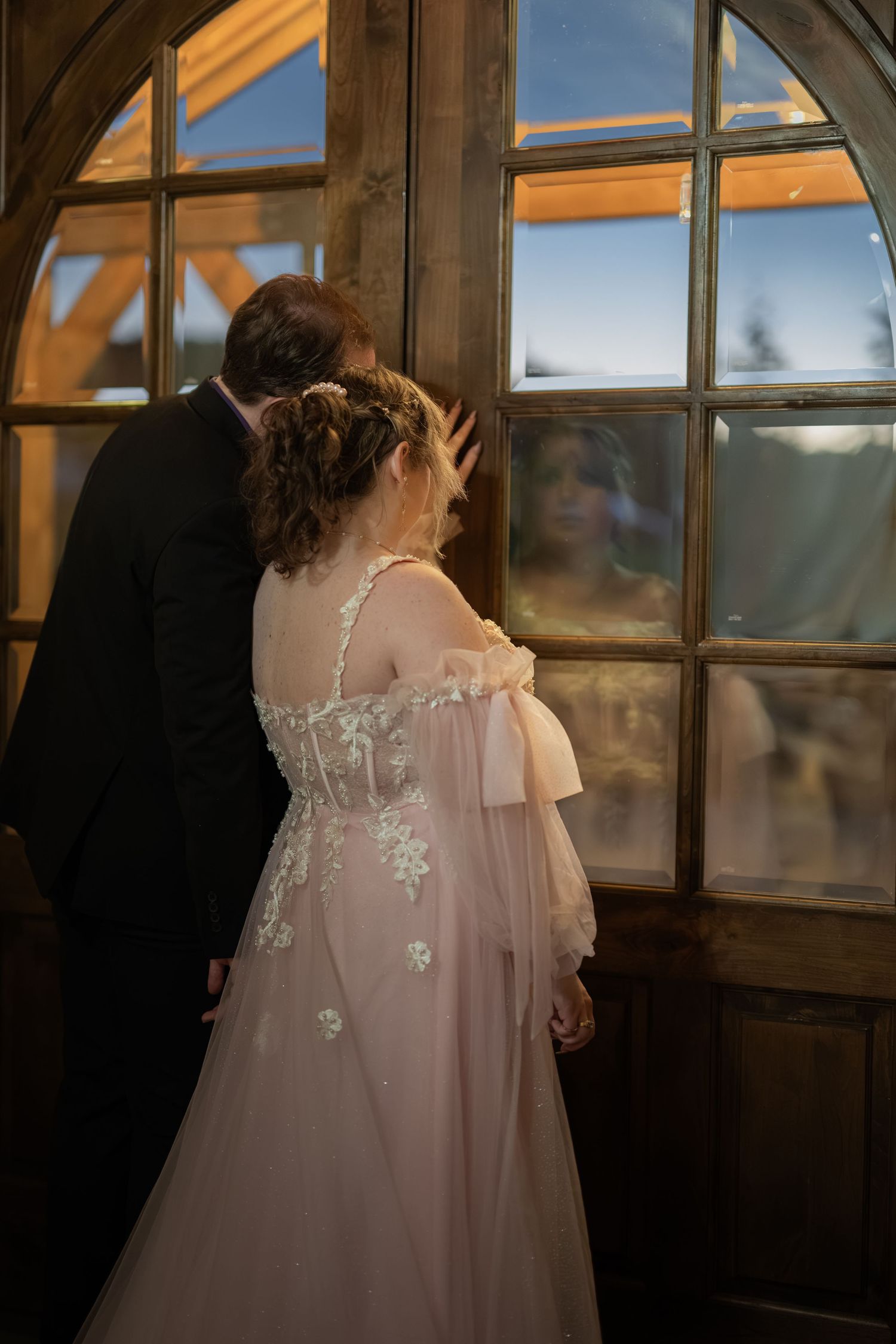 Bride in floral lace gown and groom in black suit look through vintage window, their reflection visible in glass.