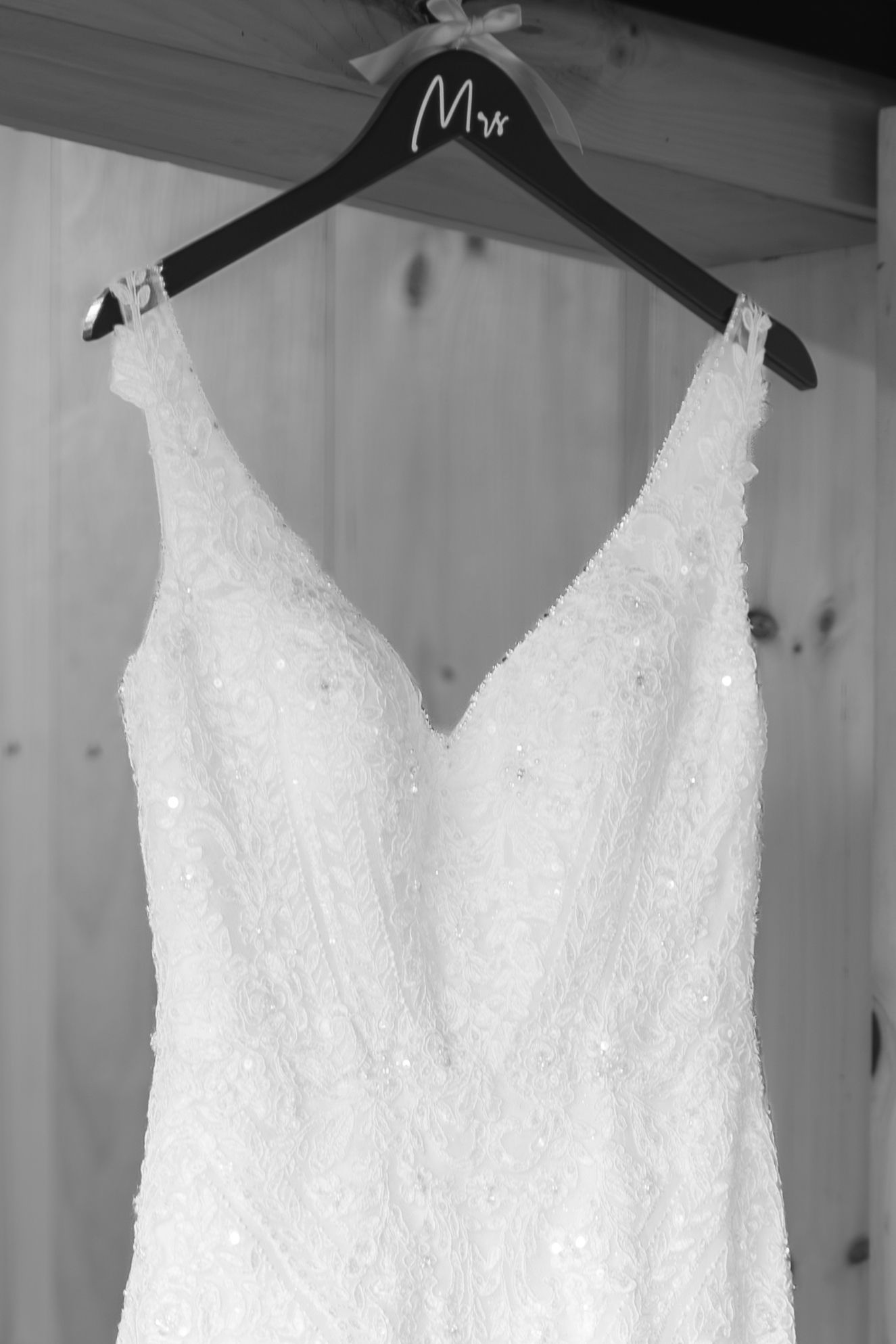 Black and white photo of a beaded lace wedding dress hanging on a wooden hanger labeled Mrs against a wood panel wall.