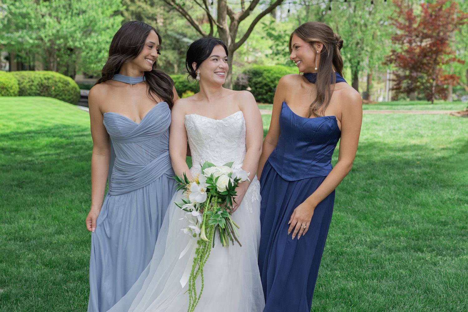 Bride in white strapless gown holding bouquet stands between two bridesmaids in blue dresses on green lawn.