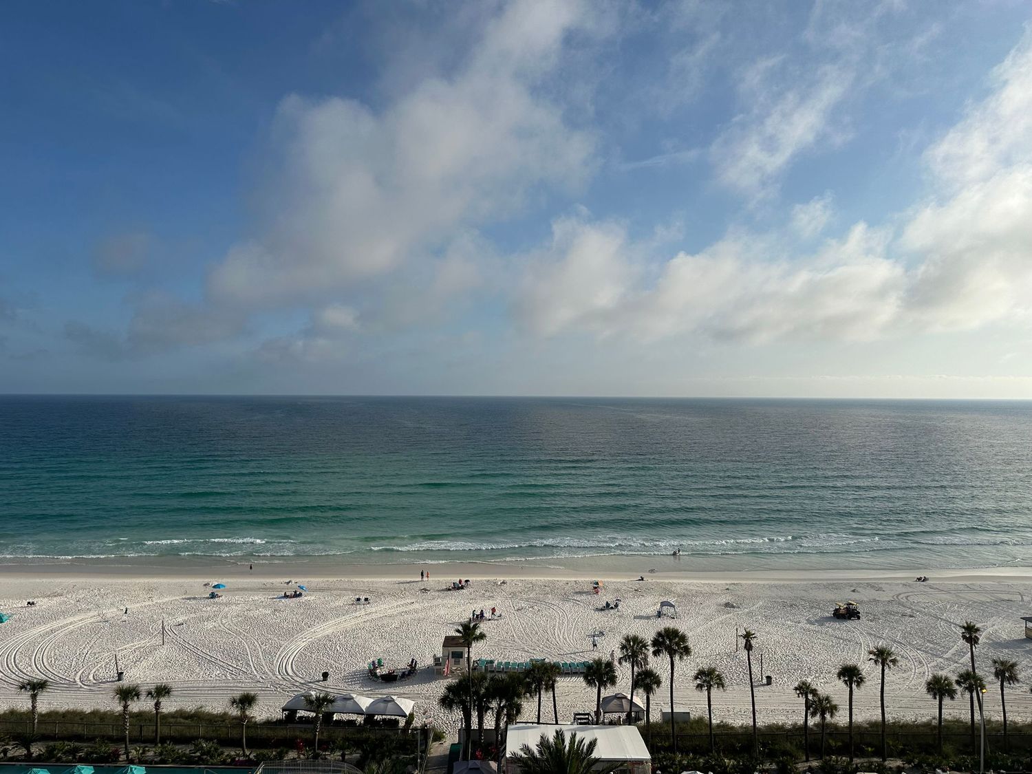 Aerial view of a pristine white sand beach with turquoise Gulf waters, palm trees, and beachgoers under a partly cloudy sky.