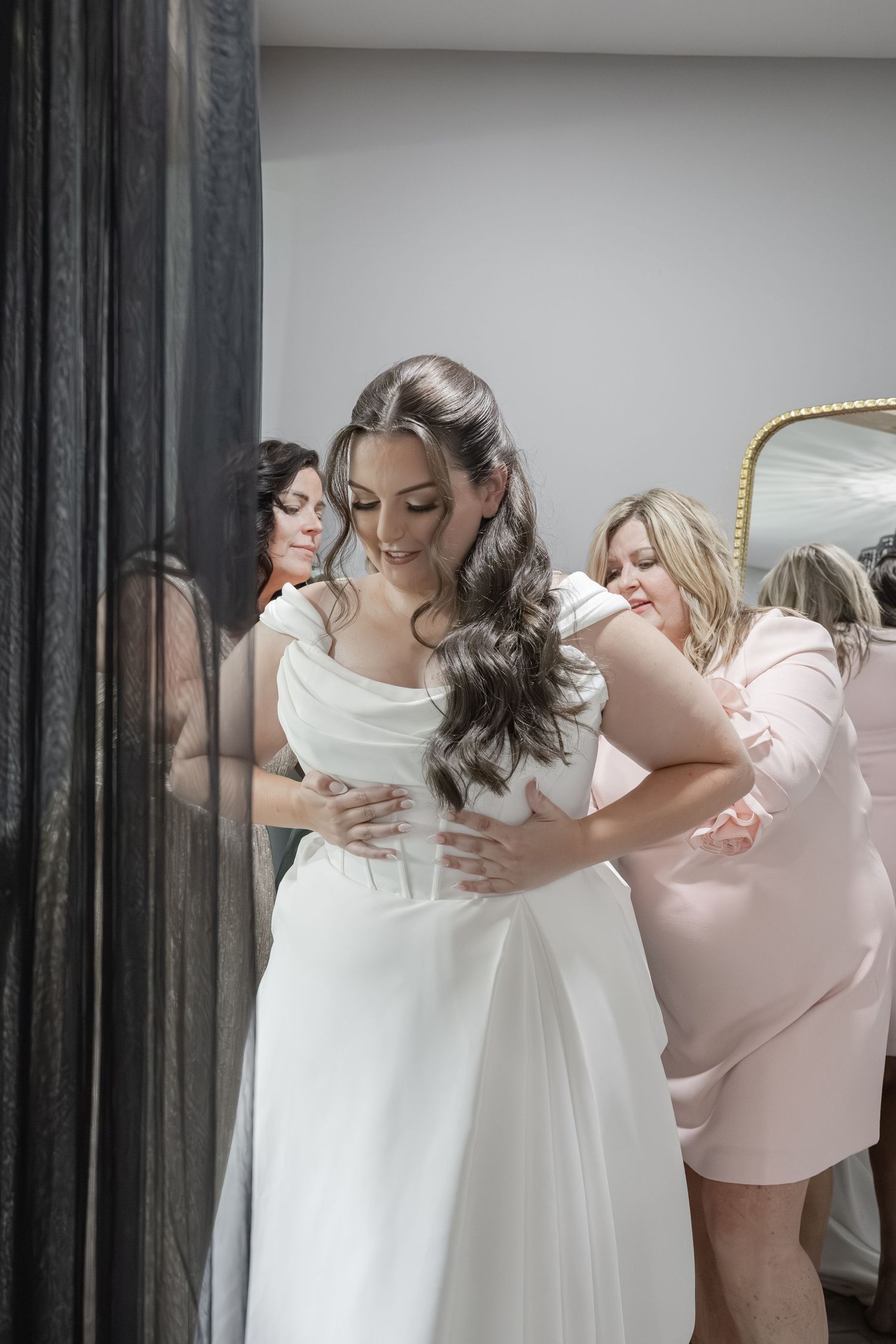 Bride in white gown being helped by two bridesmaids in pink dresses while getting ready for wedding.