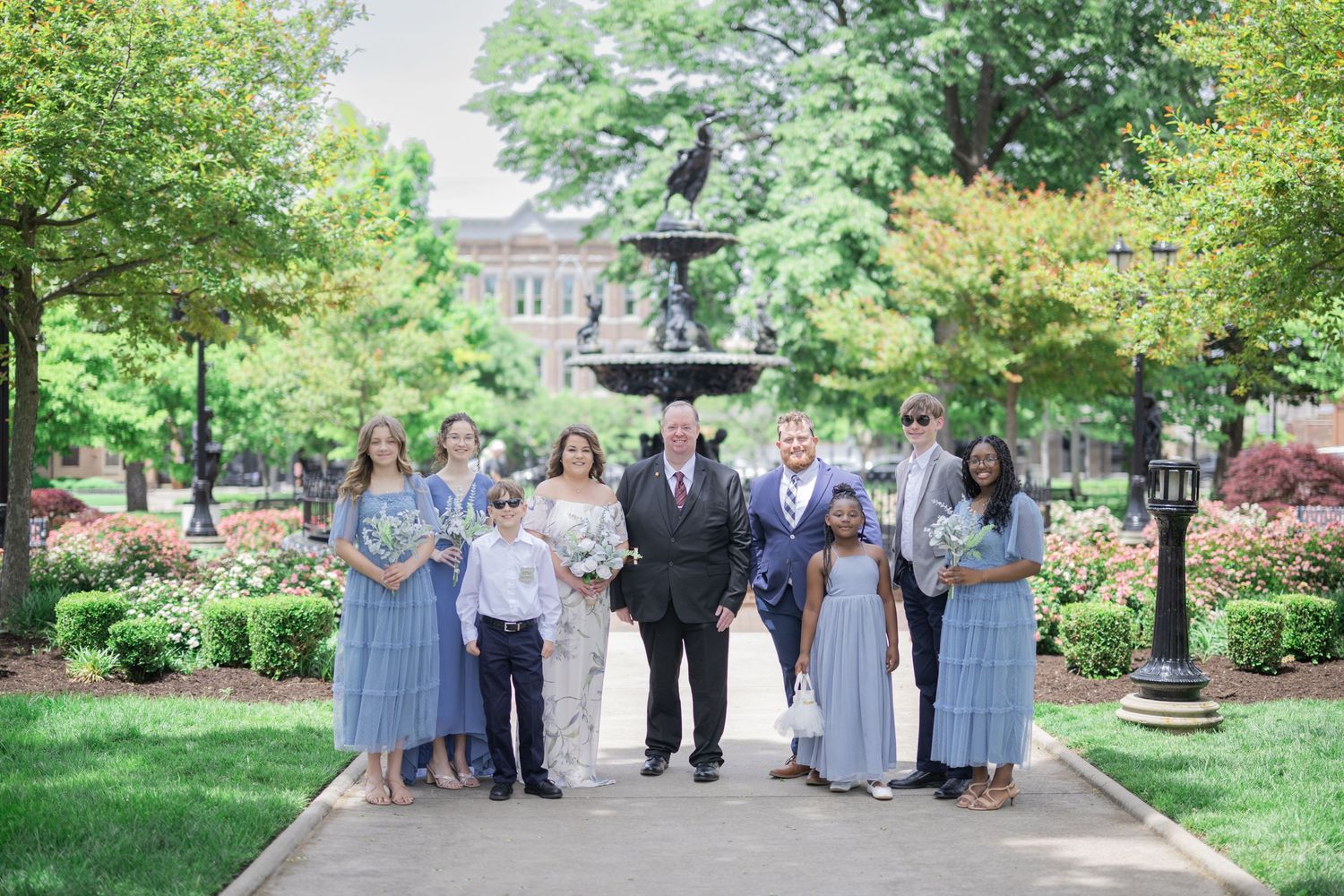 Wedding party poses in front of a fountain in a lush garden, bridesmaids in blue dresses flanking the bride and groom.