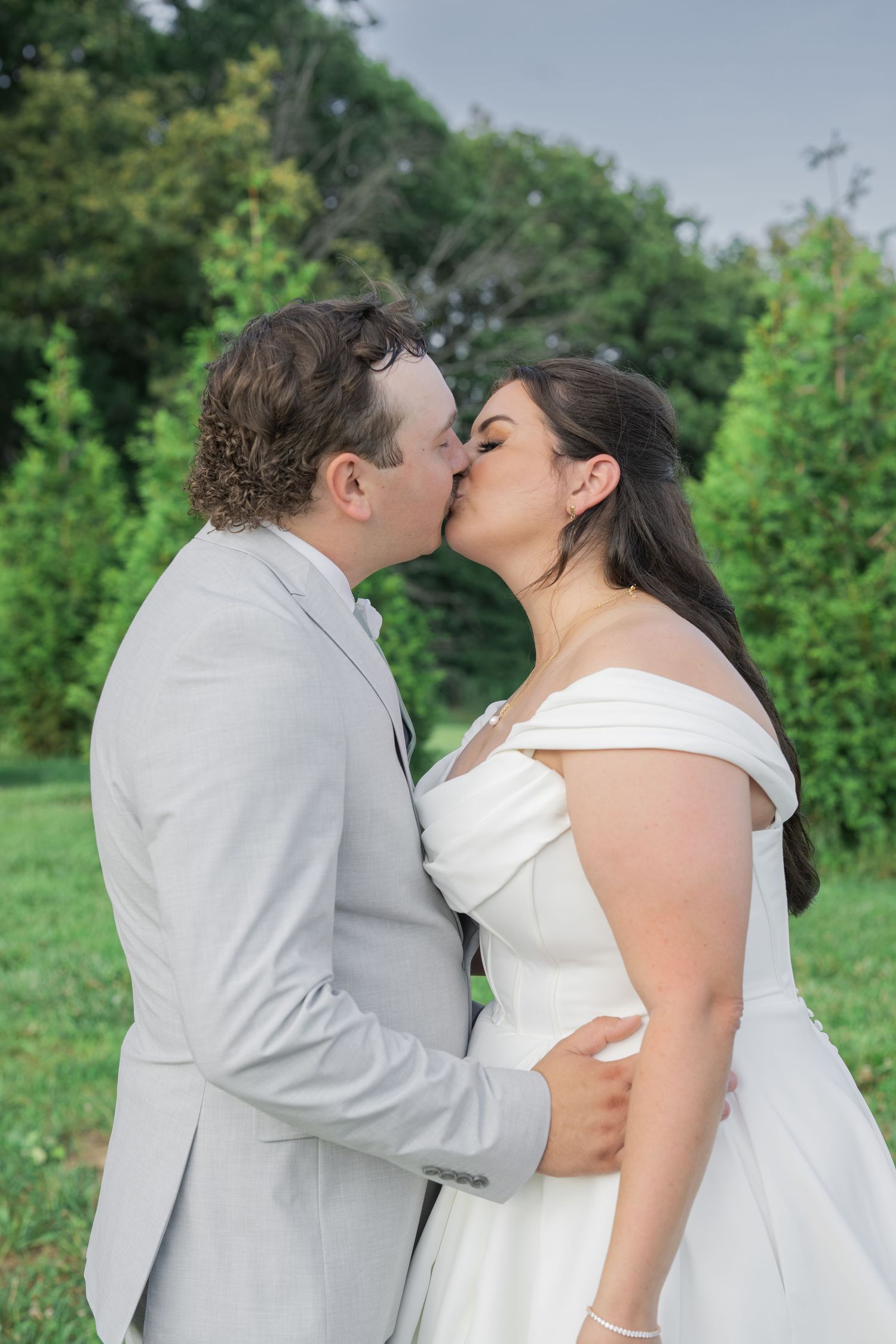 A bride and groom share a romantic kiss outdoors, dressed in a white gown and gray suit, surrounded by lush greenery.