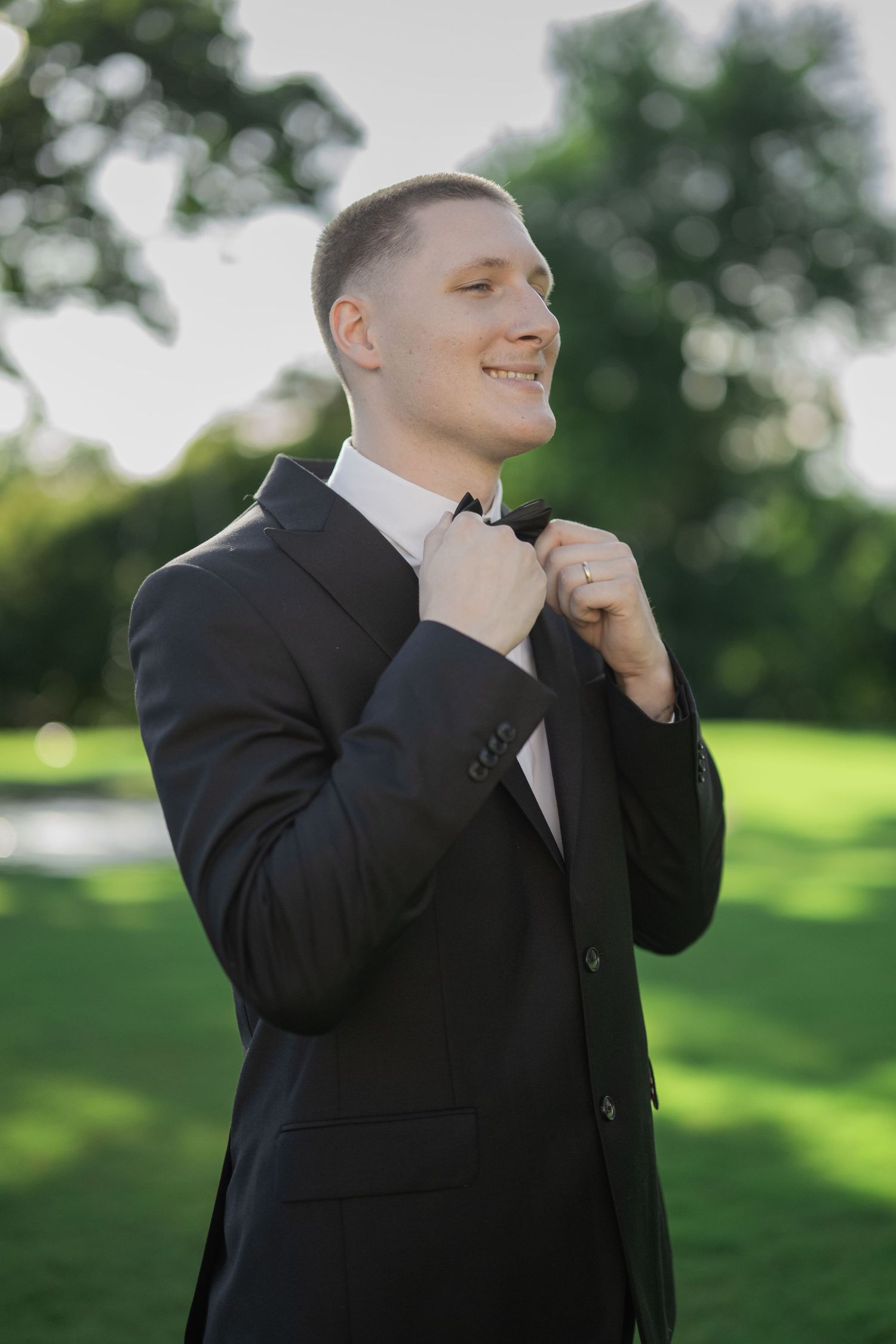 Smiling groom in black suit adjusting bow tie outdoors in green park on sunny day.