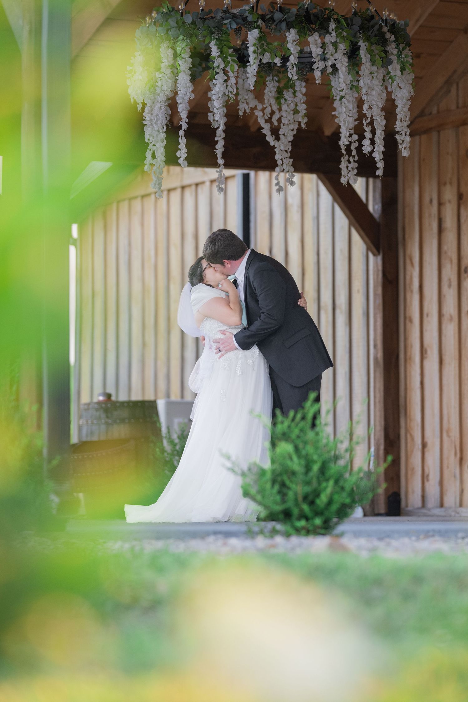 Bride and groom sharing a romantic kiss under floral decorations at a rustic wooden barn wedding venue.
