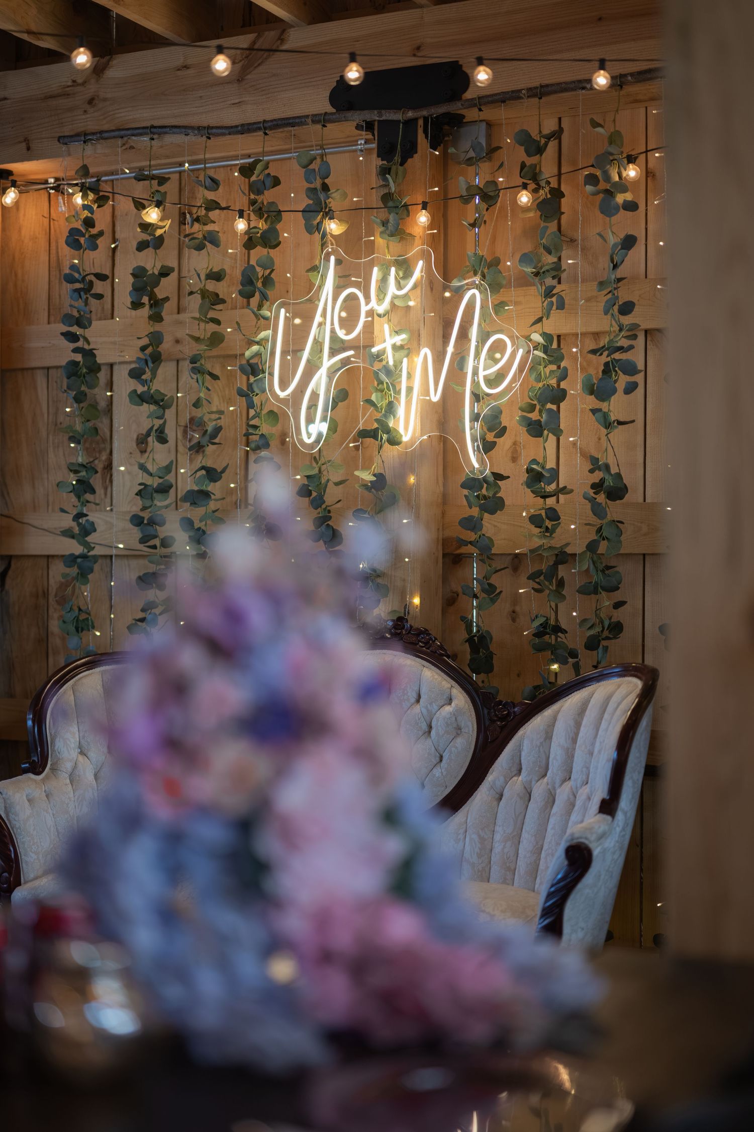Romantic wedding backdrop with neon You & Me sign, fairy lights, greenery, elegant white chairs, and floral arrangement.
