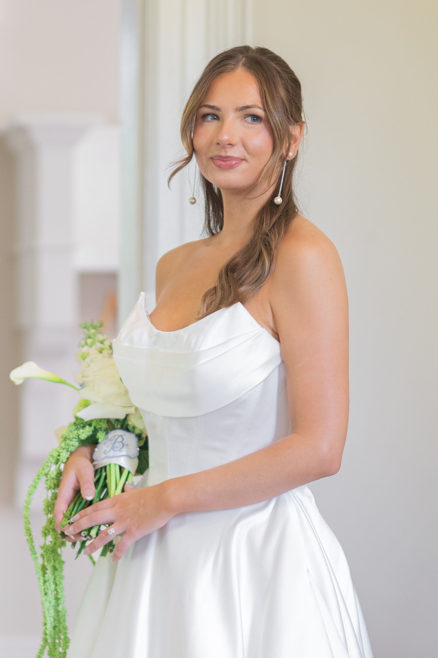 Elegant bride in a white strapless satin gown holding a green and white floral bouquet, posing indoors.