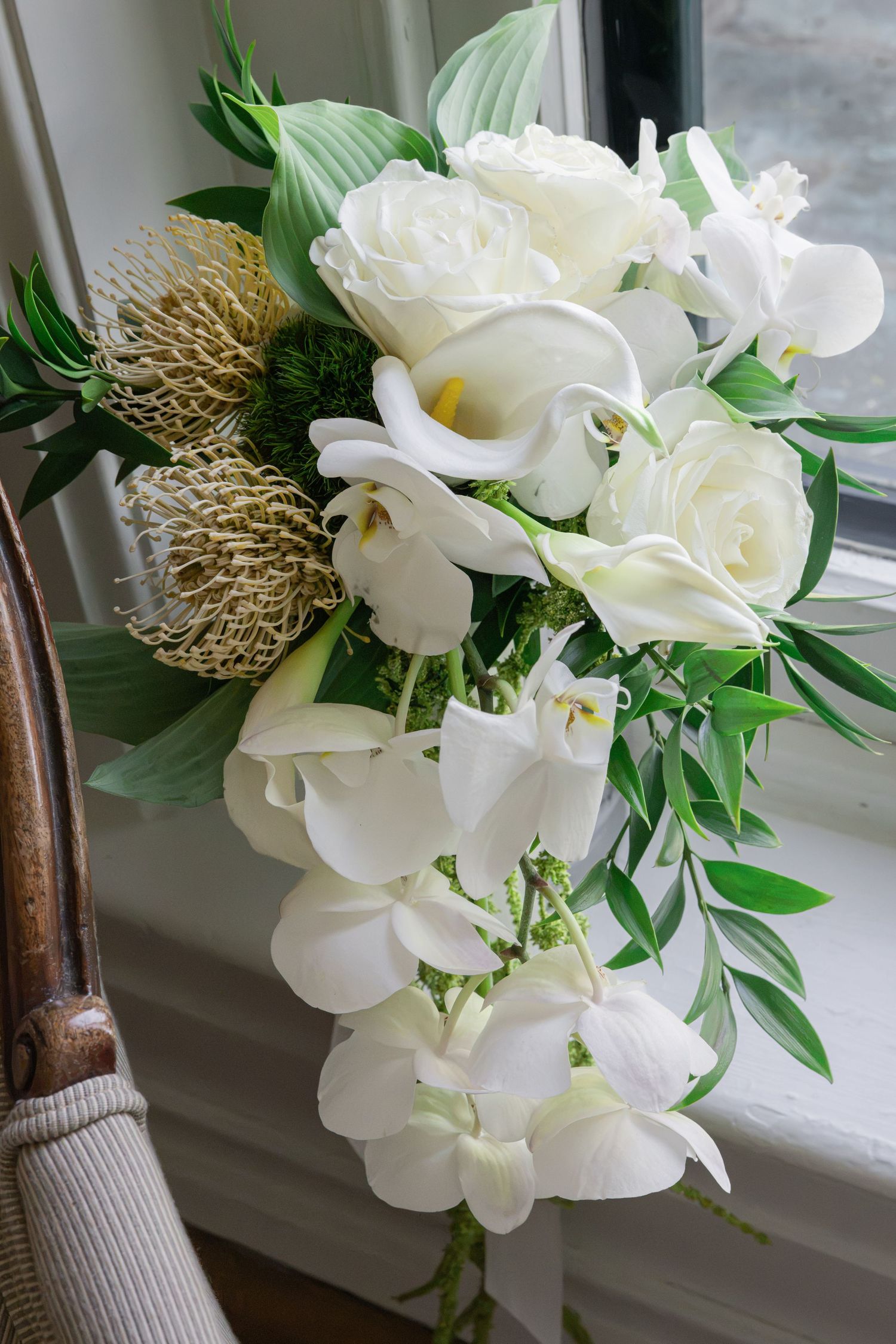 Elegant cascading bridal bouquet with white roses, calla lilies, orchids, and tropical greenery resting on a windowsill.