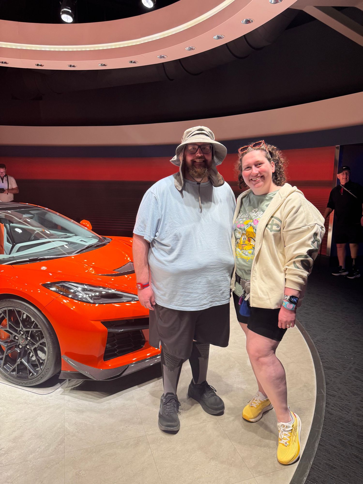 Two people posing together in front of a bright orange sports car at an auto show display.