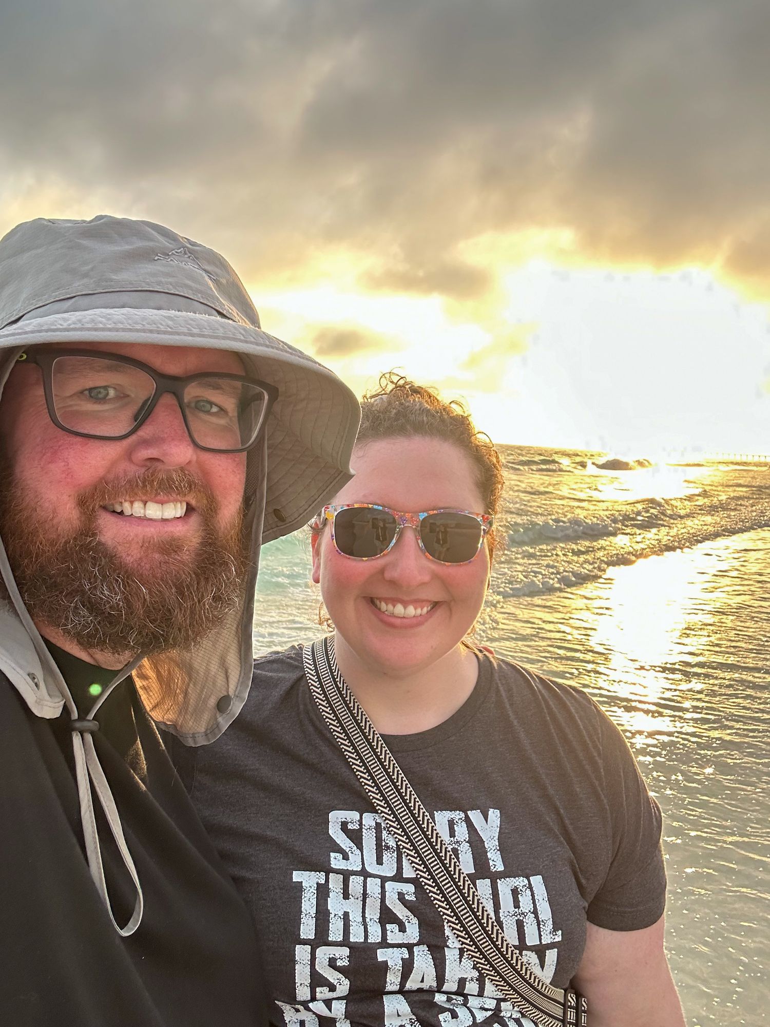 Couple smiling at sunset on a tropical beach, man in bucket hat and glasses, woman in graphic tee with sunglasses.