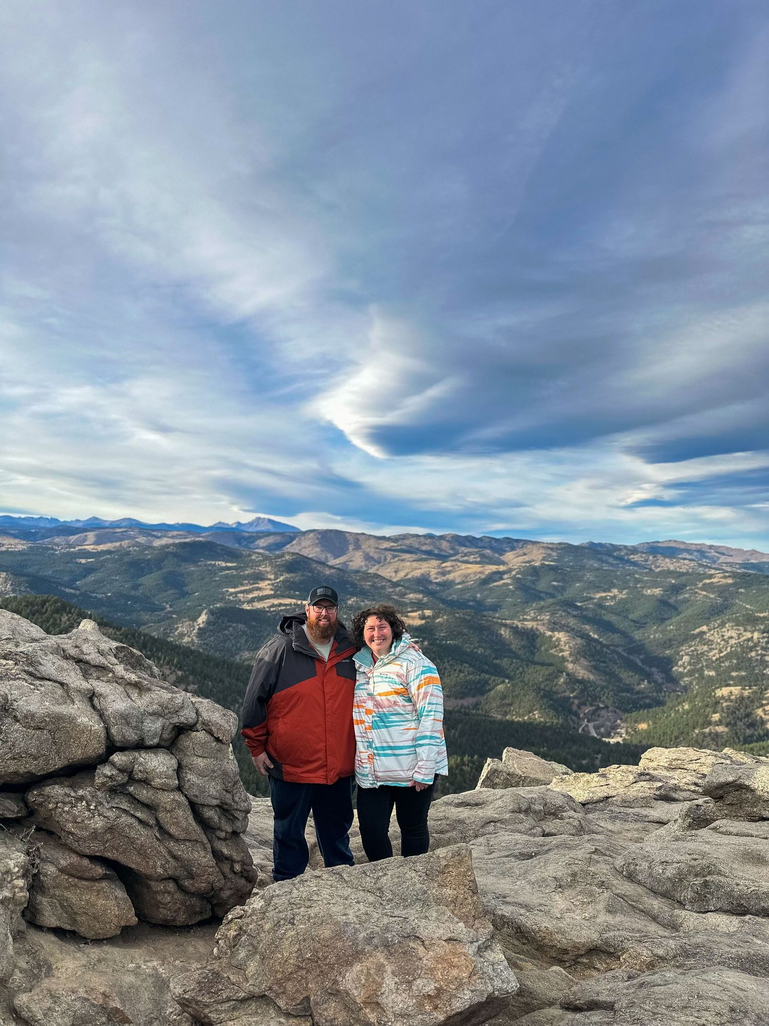 Couple posing on rocky mountain summit with sweeping valley and dramatic cloudy sky views in Colorado.