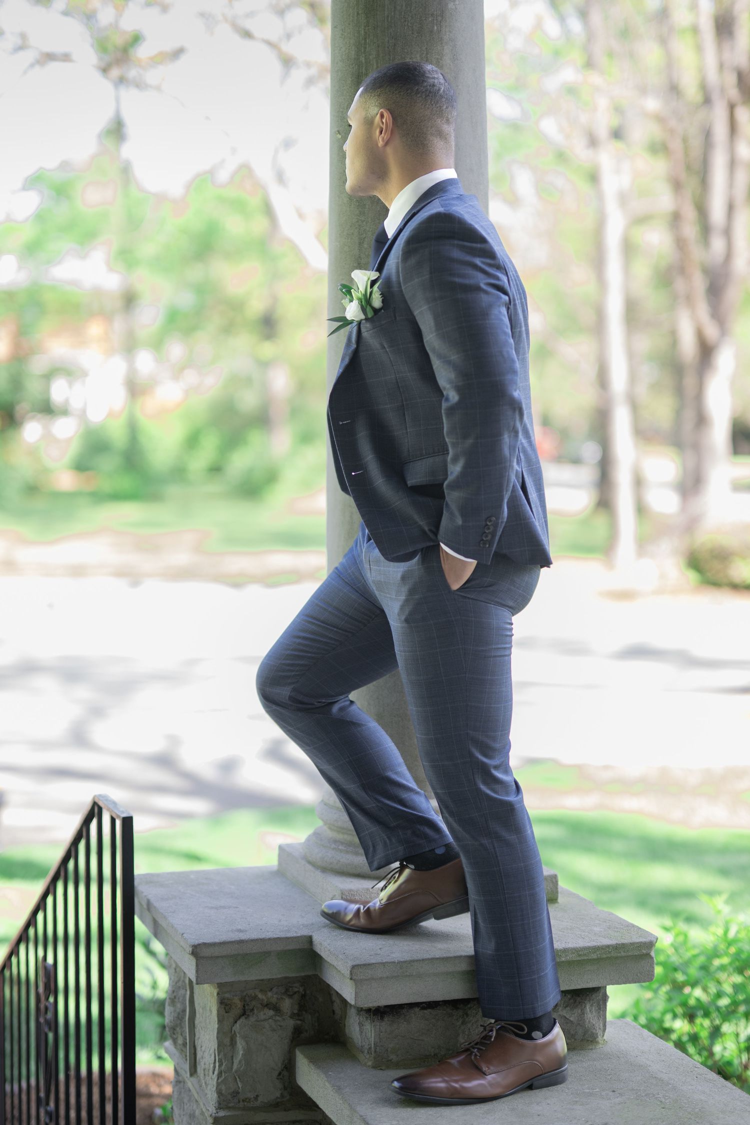Groom in navy blue suit with boutonniere leaning against stone column on outdoor steps at wedding venue.
