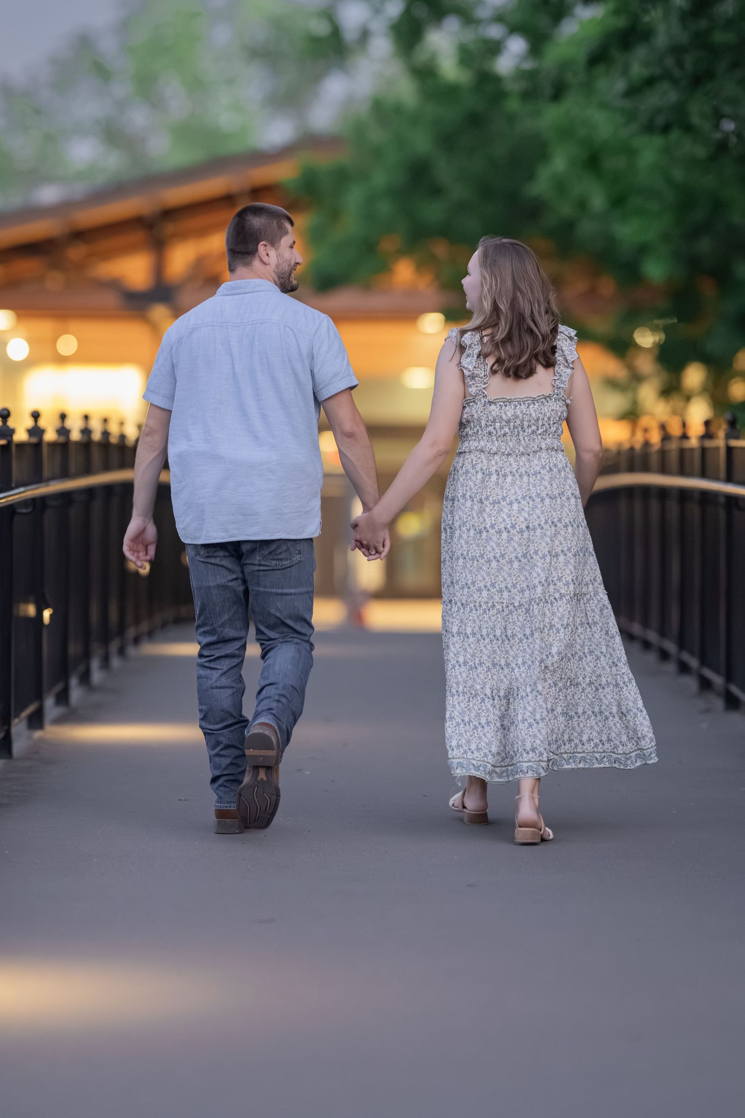 A couple holds hands while walking along a bridge at dusk, man in blue shirt and jeans, woman in floral maxi dress.