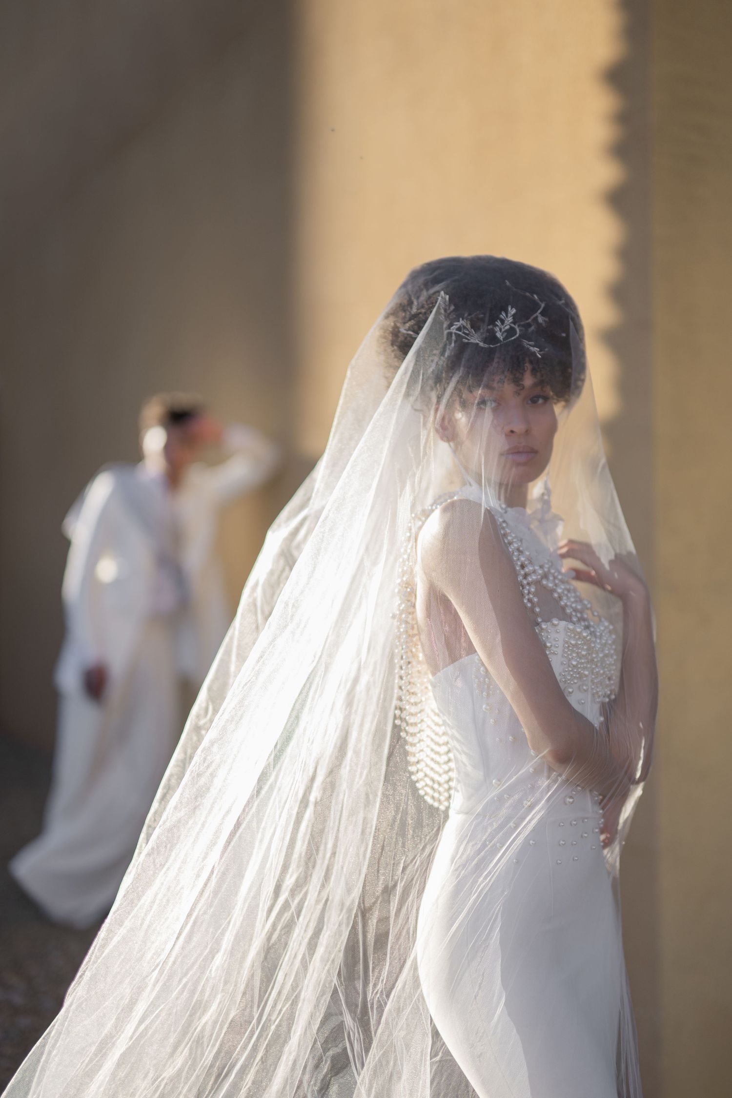 Bride in white lace gown and long veil with pearl necklace poses elegantly, groom visible in soft background.