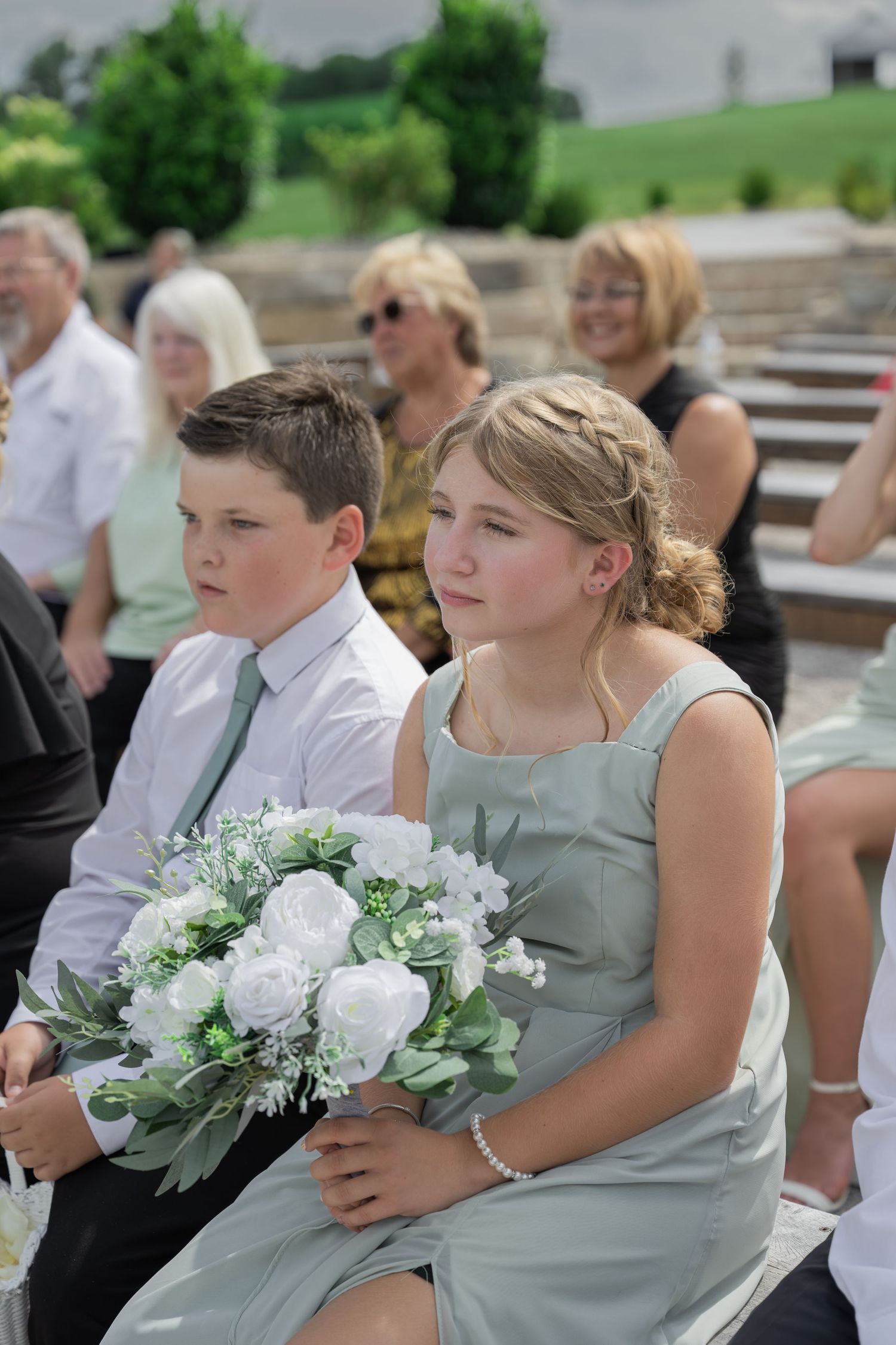 Young bridesmaid in sage green dress holds white bouquet beside boy in tie at outdoor wedding ceremony.