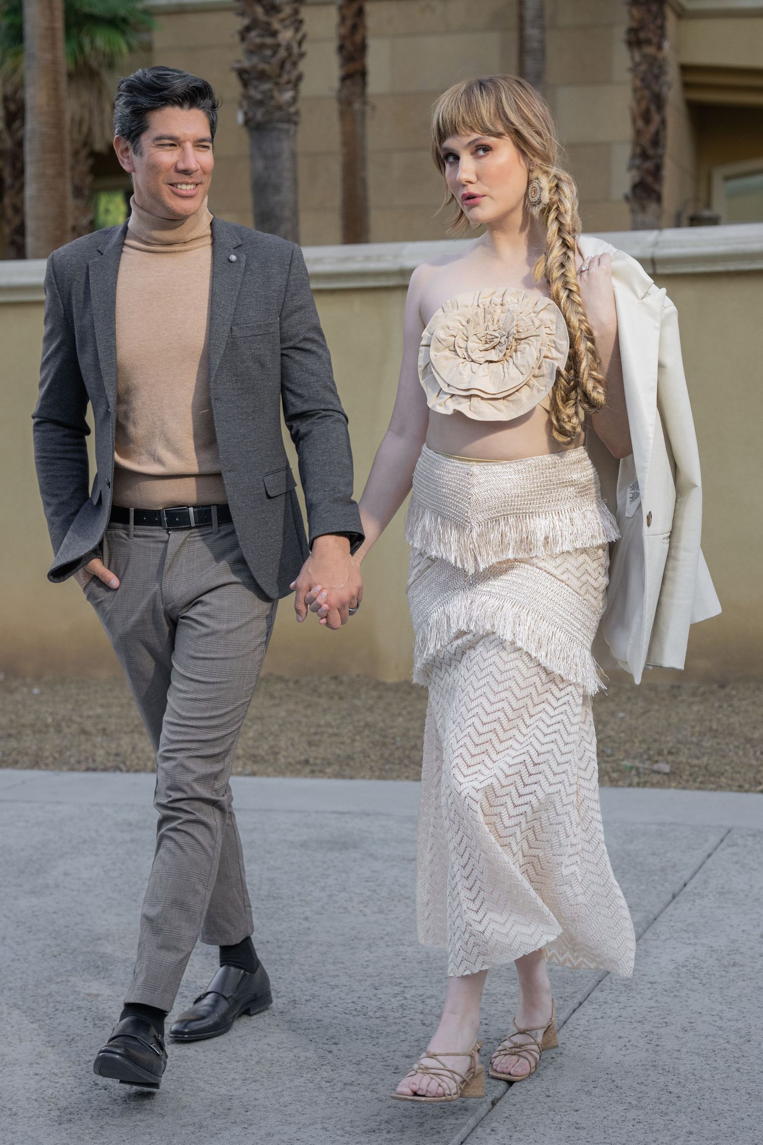 Elegant couple walking hand in hand, woman in white fringe crop top and skirt, man in gray blazer.