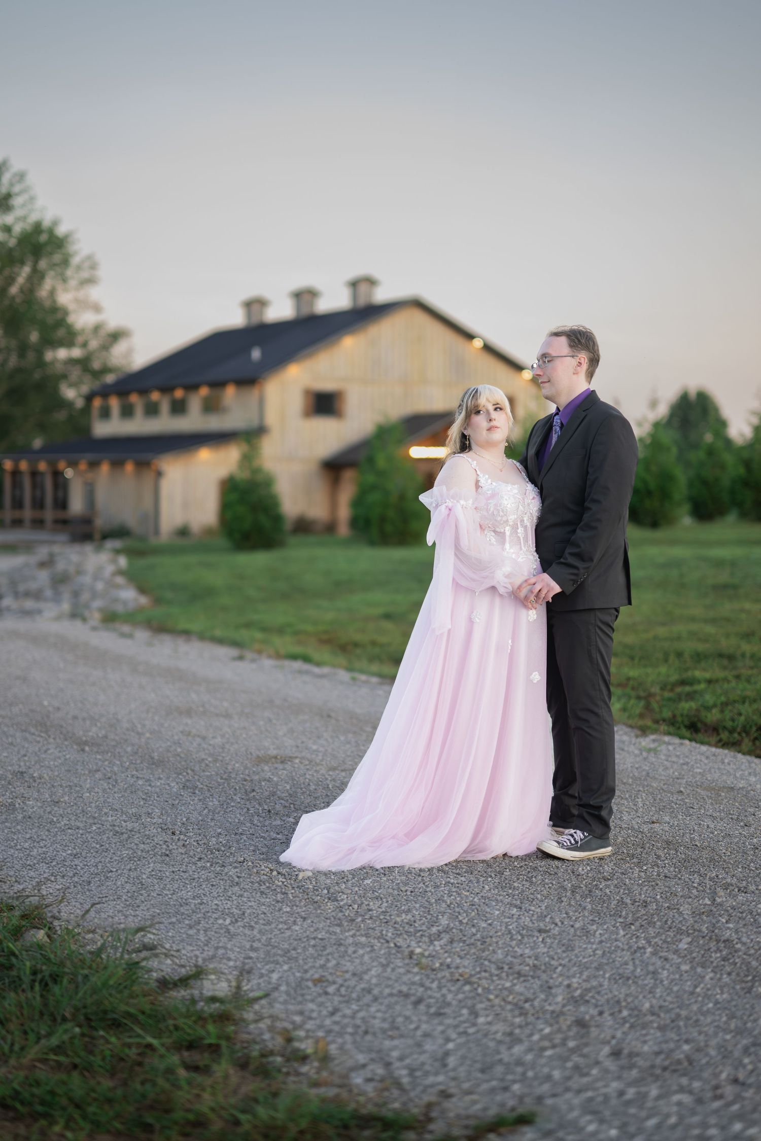 Couple in formal wear stand on gravel path before rustic venue at dusk.