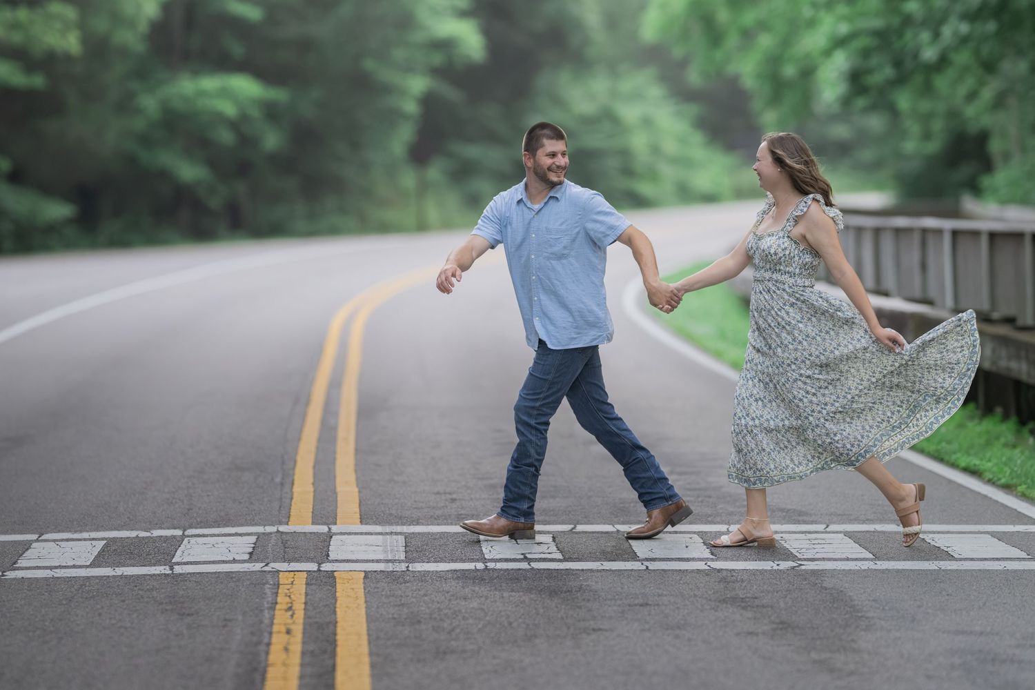 Couple holding hands crossing a road surrounded by lush green trees, woman in floral dress, man in blue shirt and jeans.
