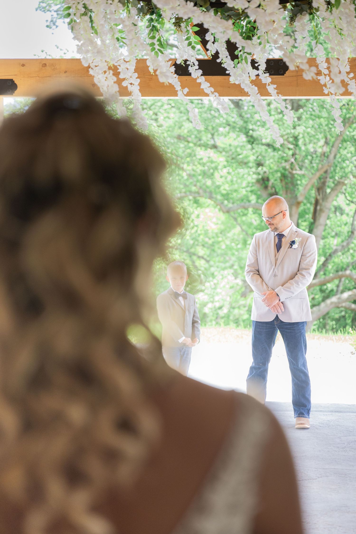 Groom in beige suit waits at altar under floral arch as bride approaches during outdoor wedding ceremony.