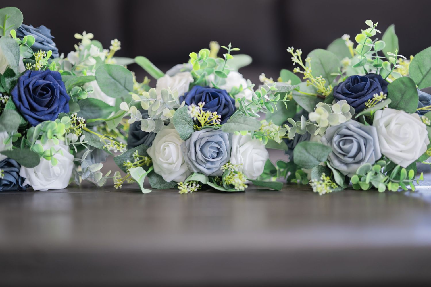 Navy blue, dusty blue, and white artificial roses with greenery arranged in elegant wedding bouquets on a gray surface.