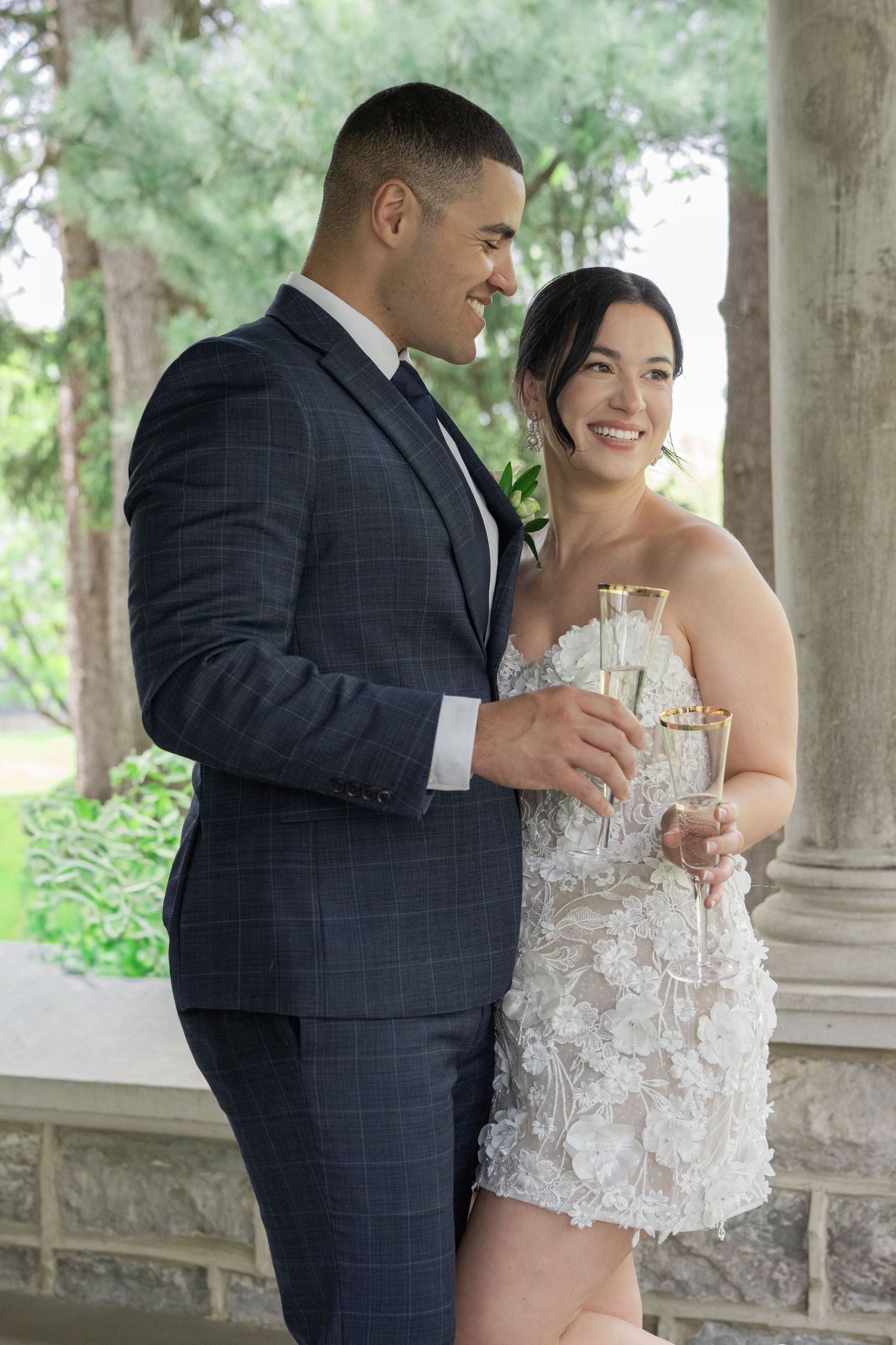 Couple in wedding attire smiling and holding champagne glasses under stone columns outdoors.