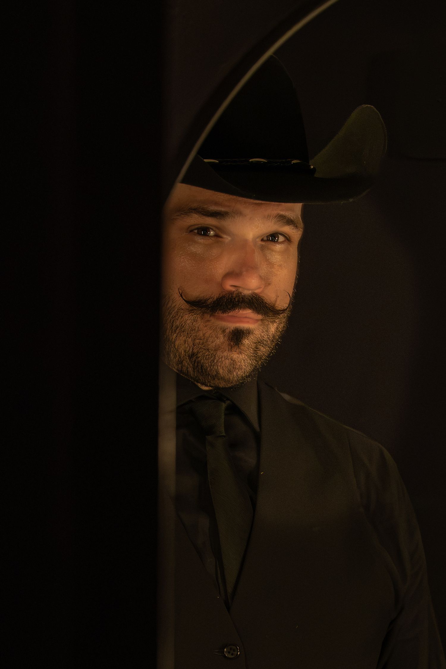 Man with handlebar mustache and black cowboy hat peering around a dark doorway in dramatic lighting.