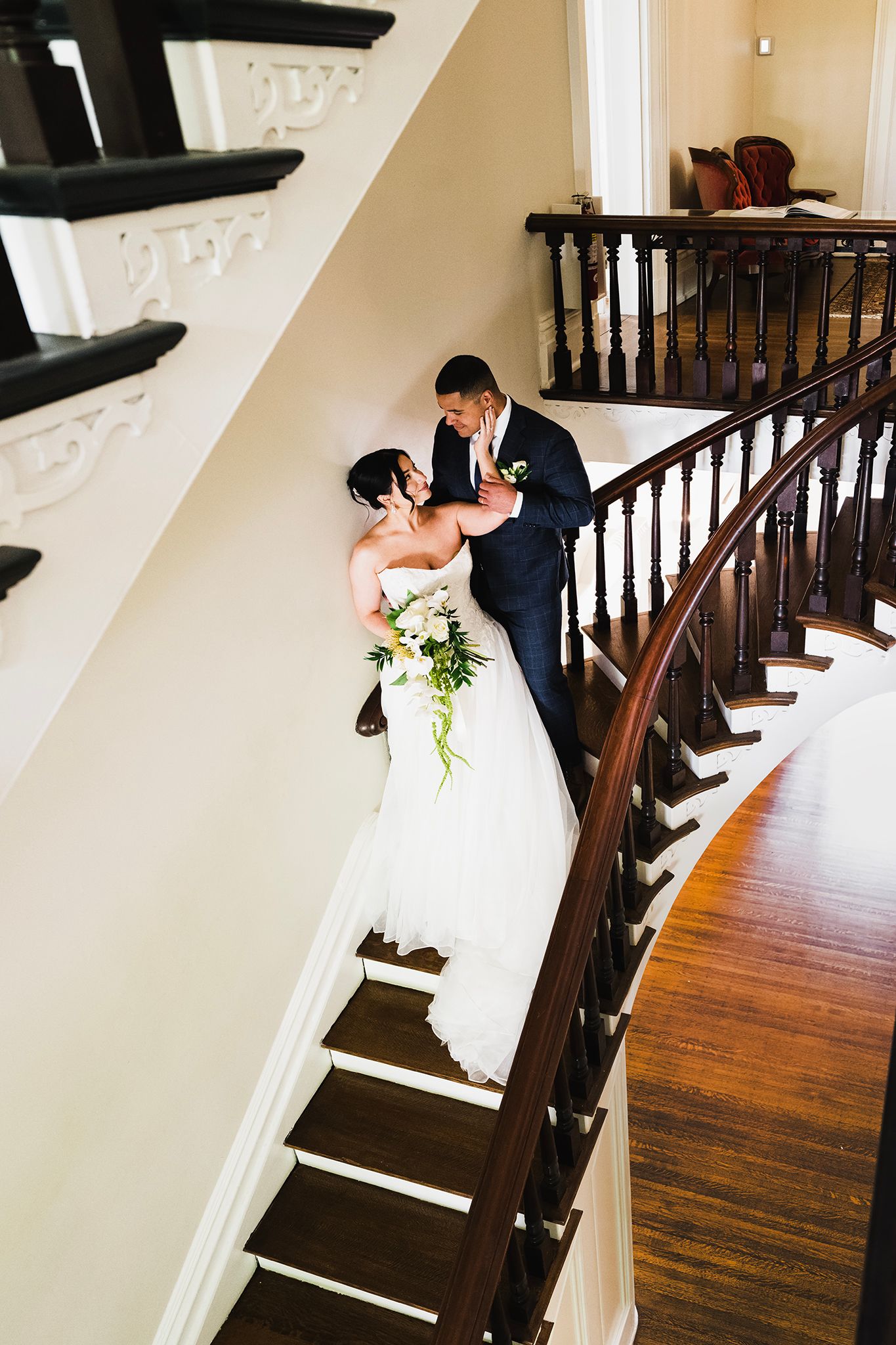 Bride and groom share a romantic moment on an elegant curved wooden staircase, bride holding a white floral bouquet.