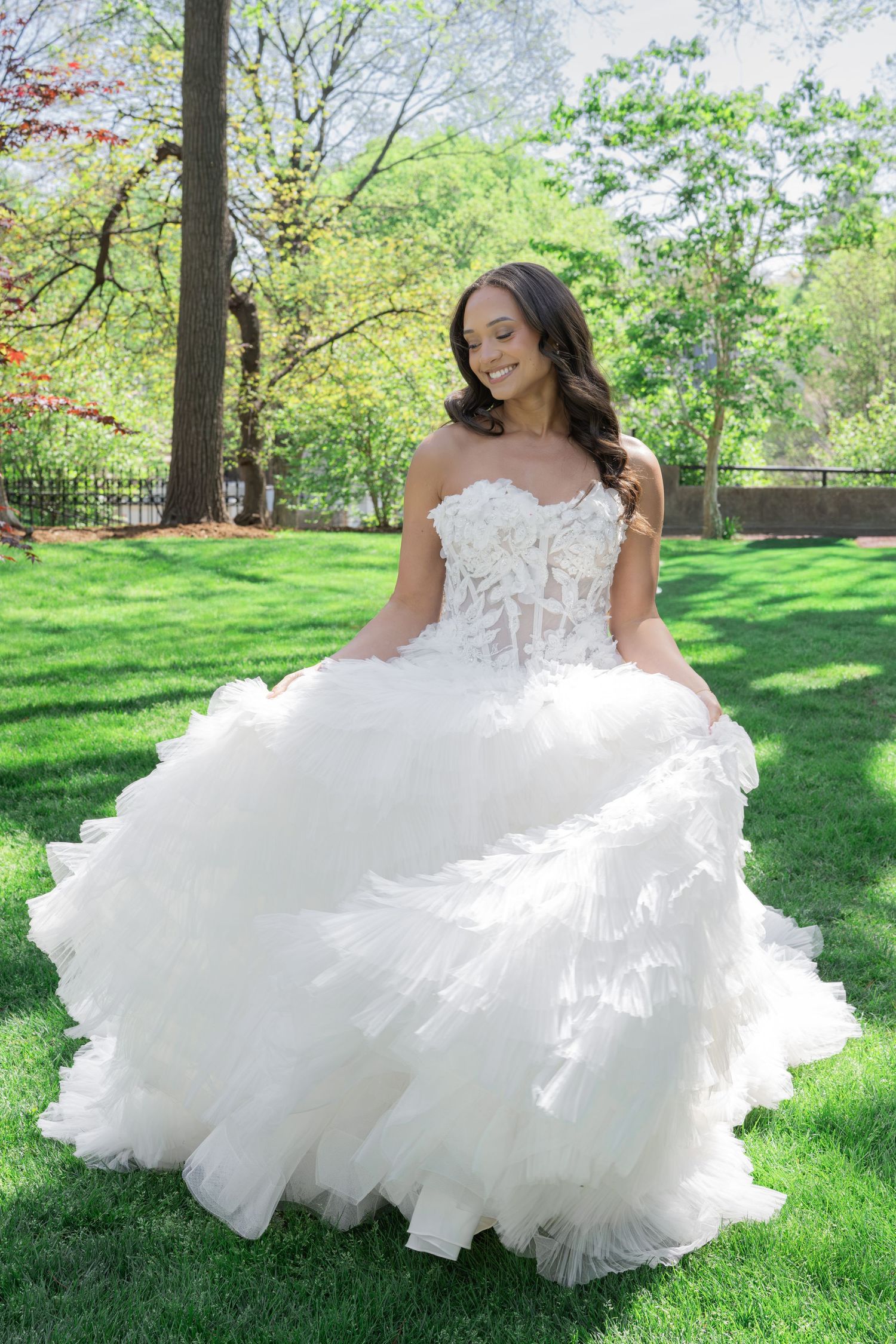 Bride in a strapless white ball gown with lace bodice and ruffled skirt posing outdoors on green grass.