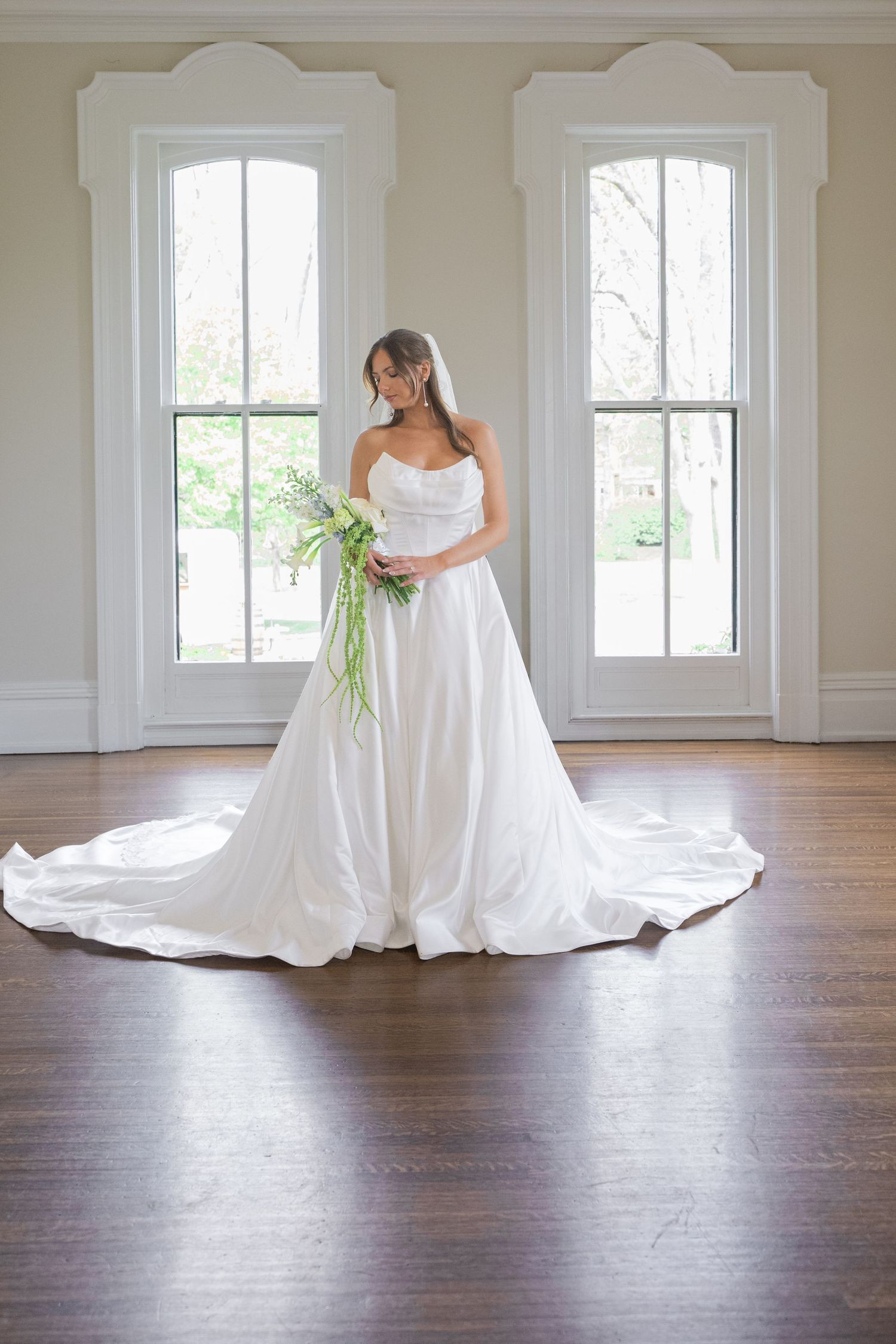 Bride in strapless white ball gown with long train holds green bouquet, standing between two windows on hardwood floor.