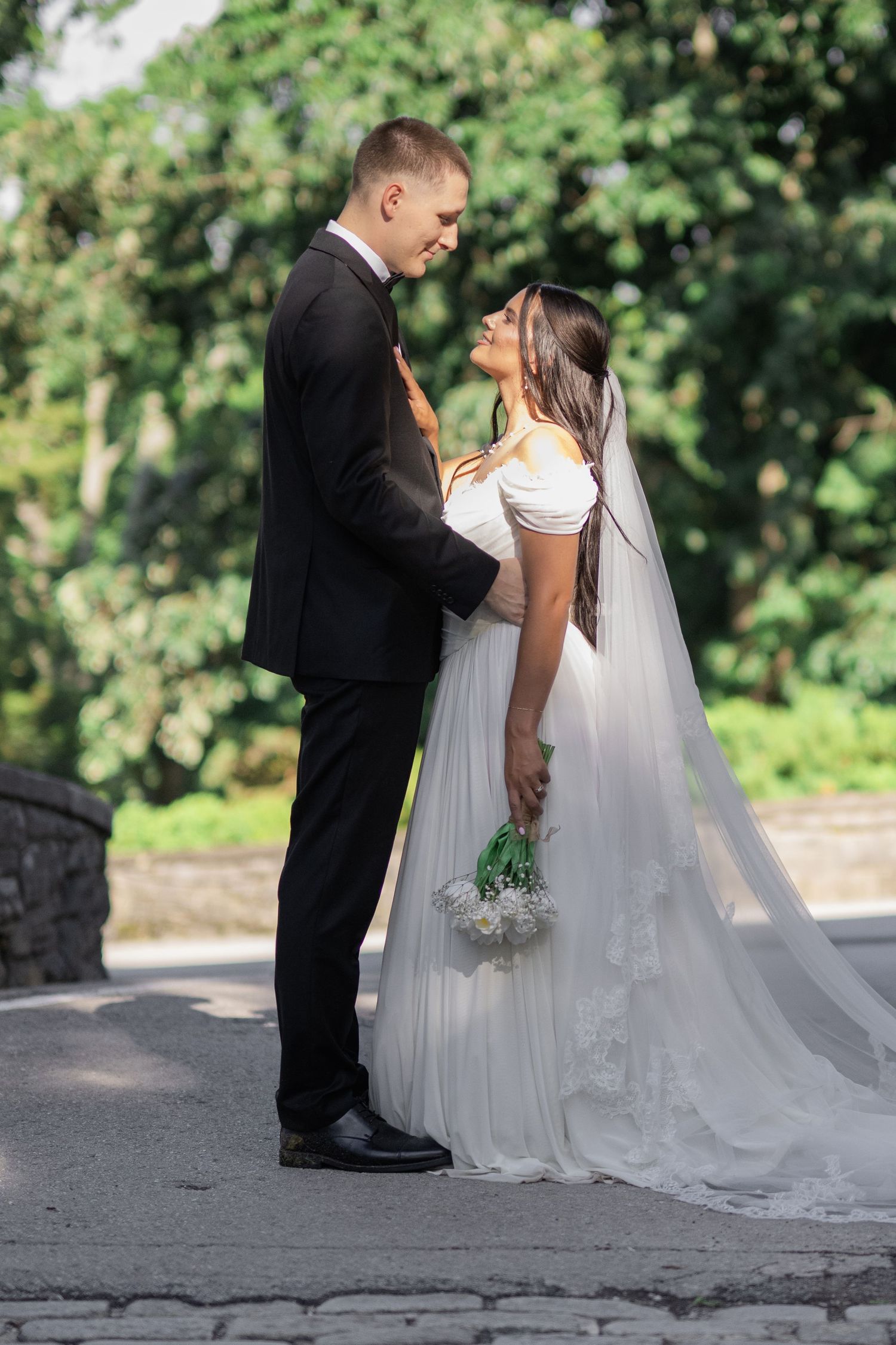 Bride and groom share a romantic moment outdoors, with the bride holding a white bouquet and wearing a flowing veil.