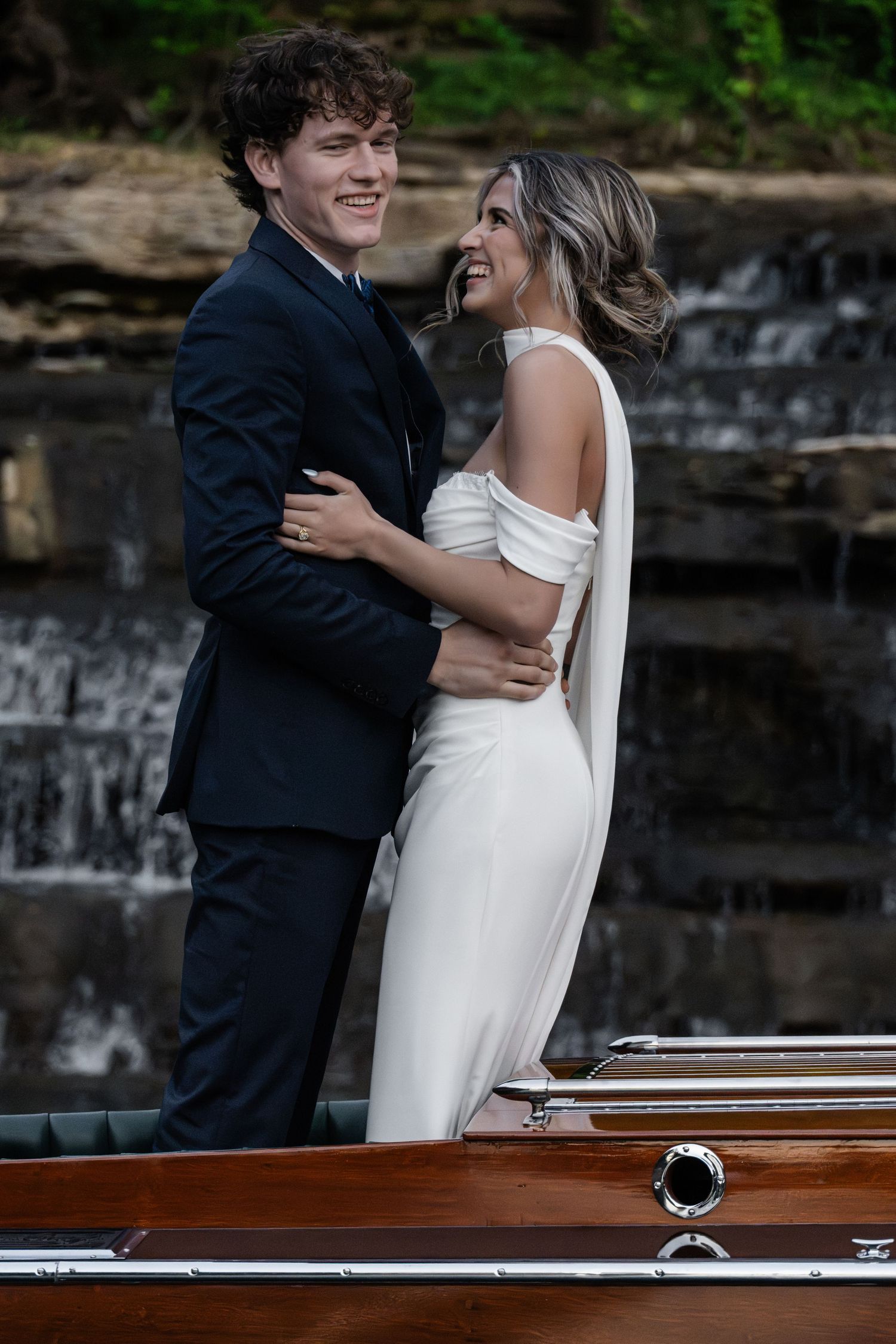 Couple in wedding attire embracing on a wooden boat with a waterfall backdrop, bride in white gown, groom in navy suit.