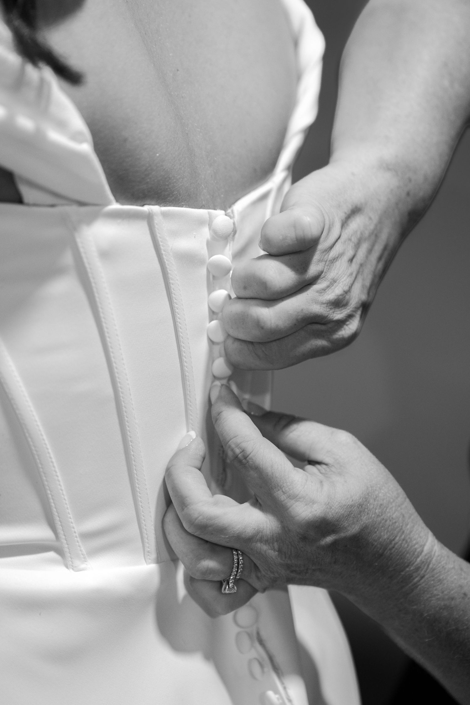 Black and white photo of hands buttoning the back of a white wedding dress corset.
