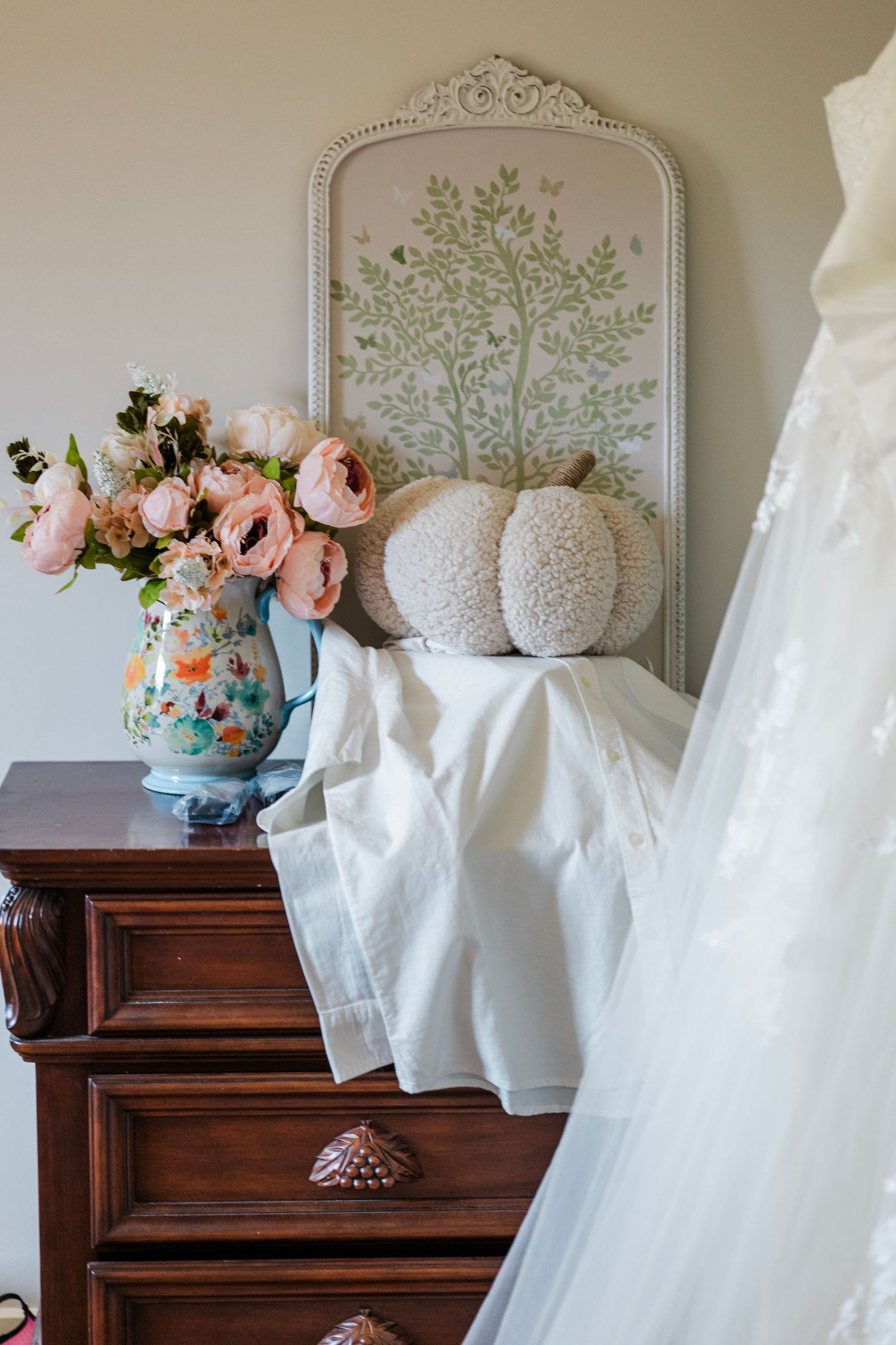 Elegant wedding scene with pink roses in floral vase on wooden dresser, decorative mirror, and white bridal gown.