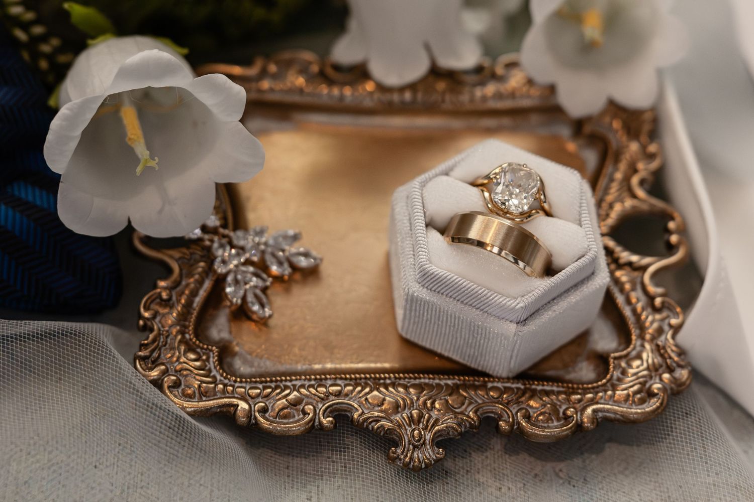 Gold wedding bands and diamond engagement ring in white velvet box on ornate brass tray with white flowers.