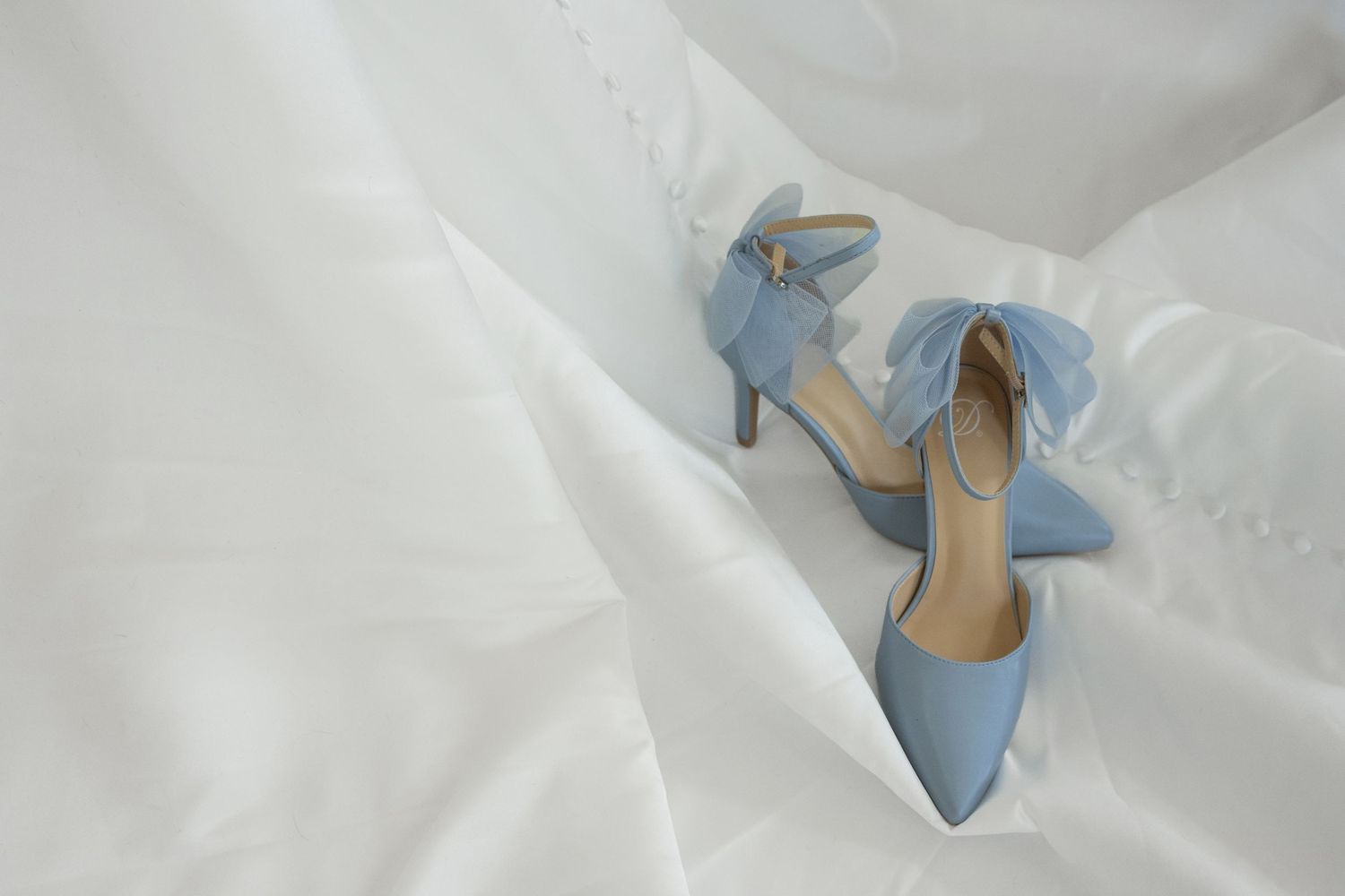 Light blue satin heels with bow detail resting on white wedding dress fabric, perfect bridal something blue shoes.