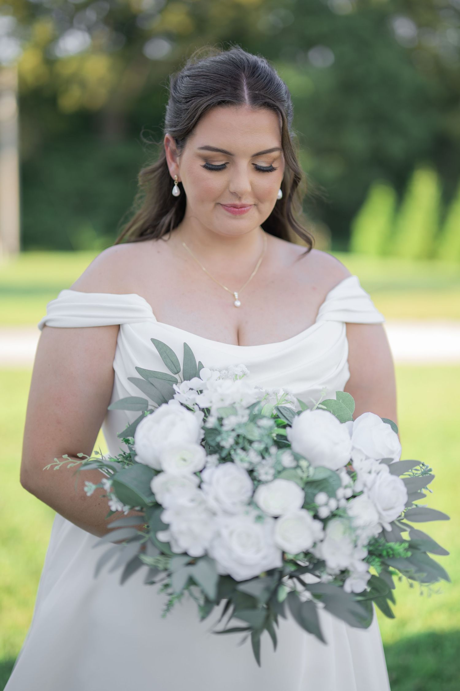 Bride in off-shoulder white gown gazes at lush white rose and greenery bouquet outdoors.