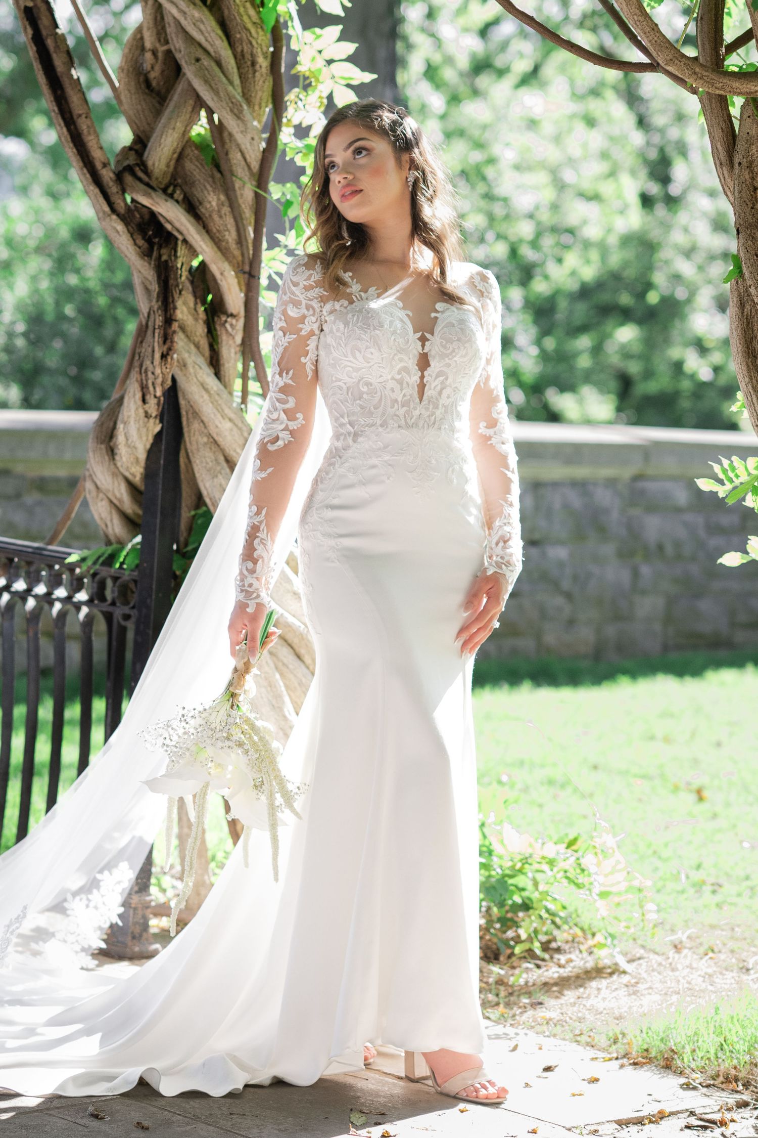 Bride in long-sleeve lace wedding gown with sheer bodice holds bouquet outdoors among lush greenery.