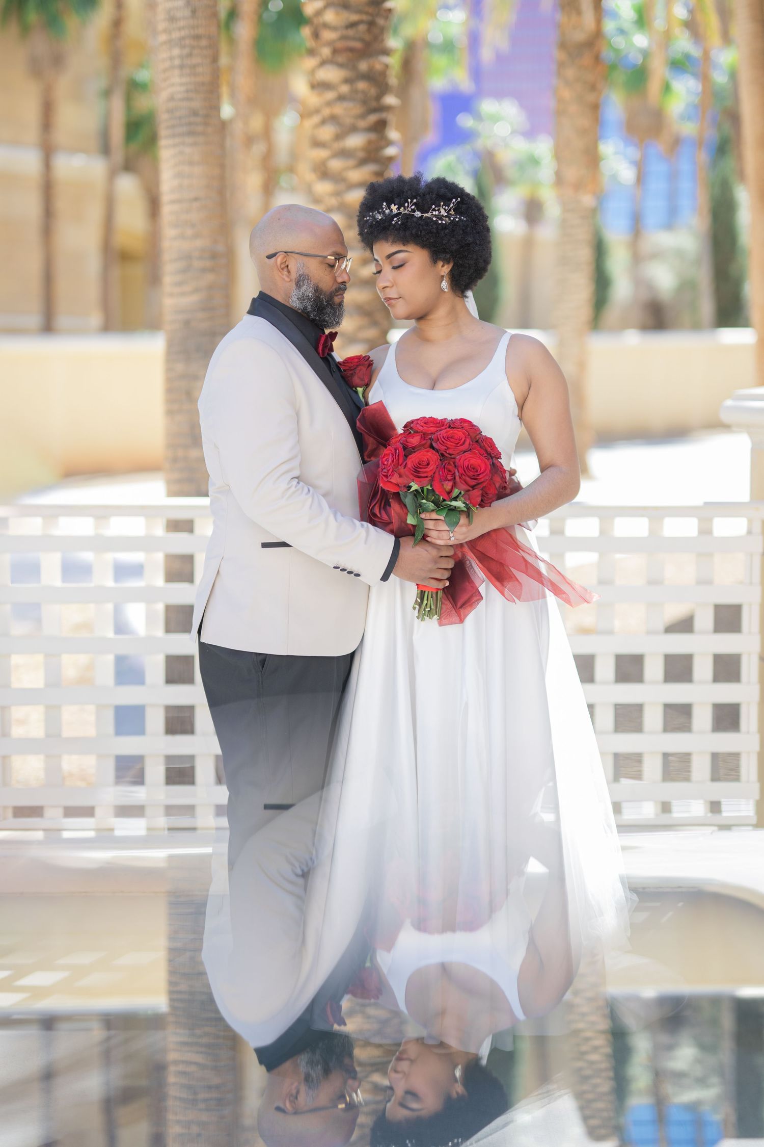 Elegant bride and groom in white wedding attire embrace holding red roses bouquet outdoors near palm trees and fountain.