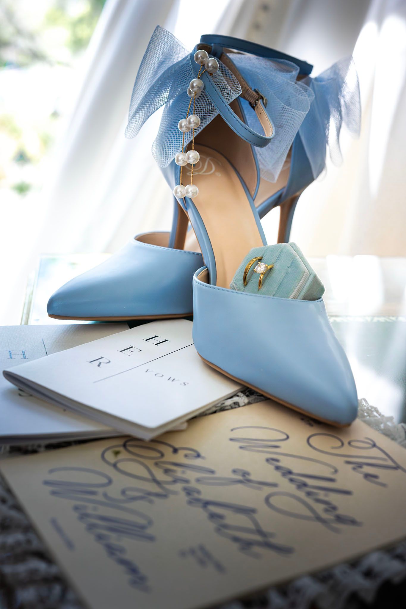 Light blue bridal heels with pearl ankle straps, tulle bow, and wedding rings resting on vow booklets.