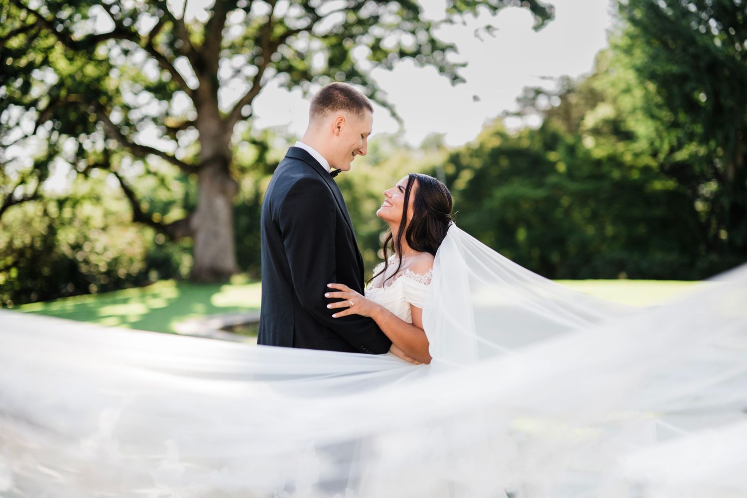 Bride and groom share a romantic moment outdoors, her veil flowing in the breeze, surrounded by lush green trees. Cheekwood, Nashville Tenn