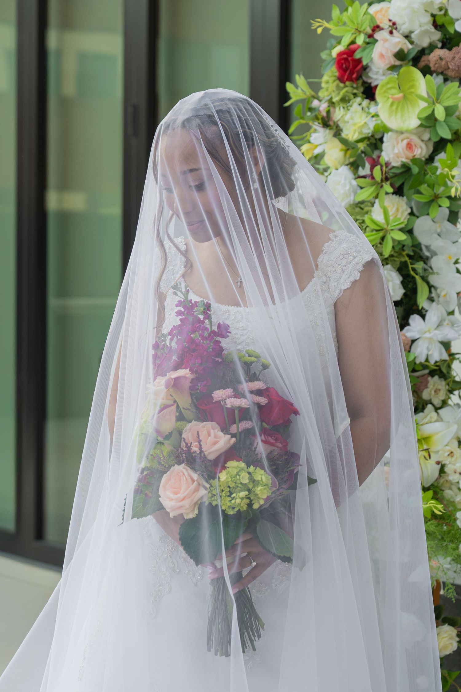 Bride in white lace dress and veil holding colorful bouquet of roses and flowers near floral arrangement.
