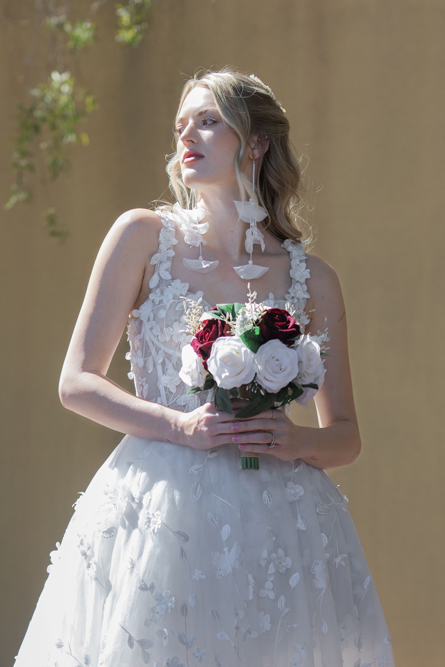 Blonde bride in white floral gown holding red and white rose bouquet, standing outdoors in sunlight.