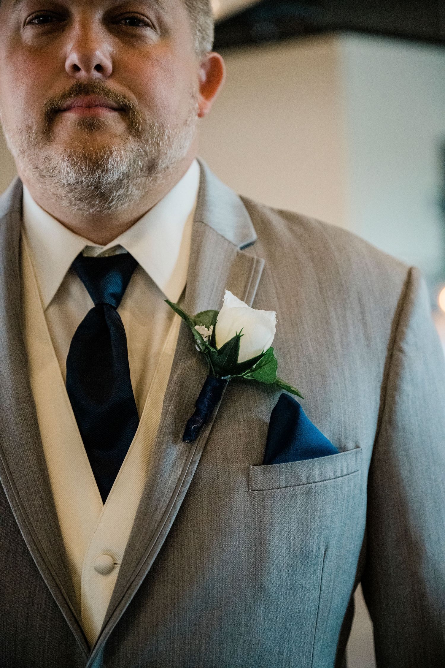 Groom in gray suit with navy tie, white rose boutonniere, and navy pocket square at wedding ceremony.