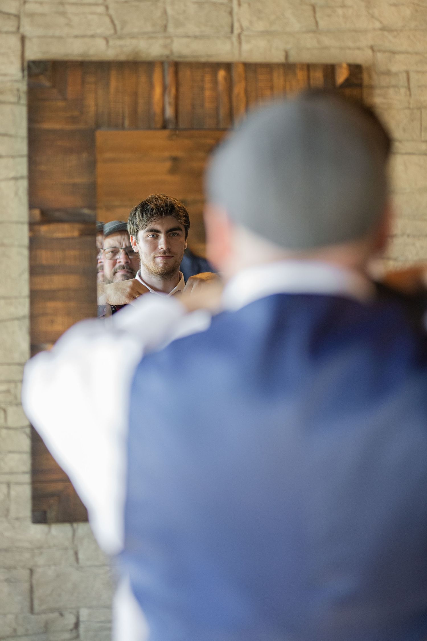 Groom adjusting his tie in a rustic wooden mirror with a reflection of a young man assisting him.