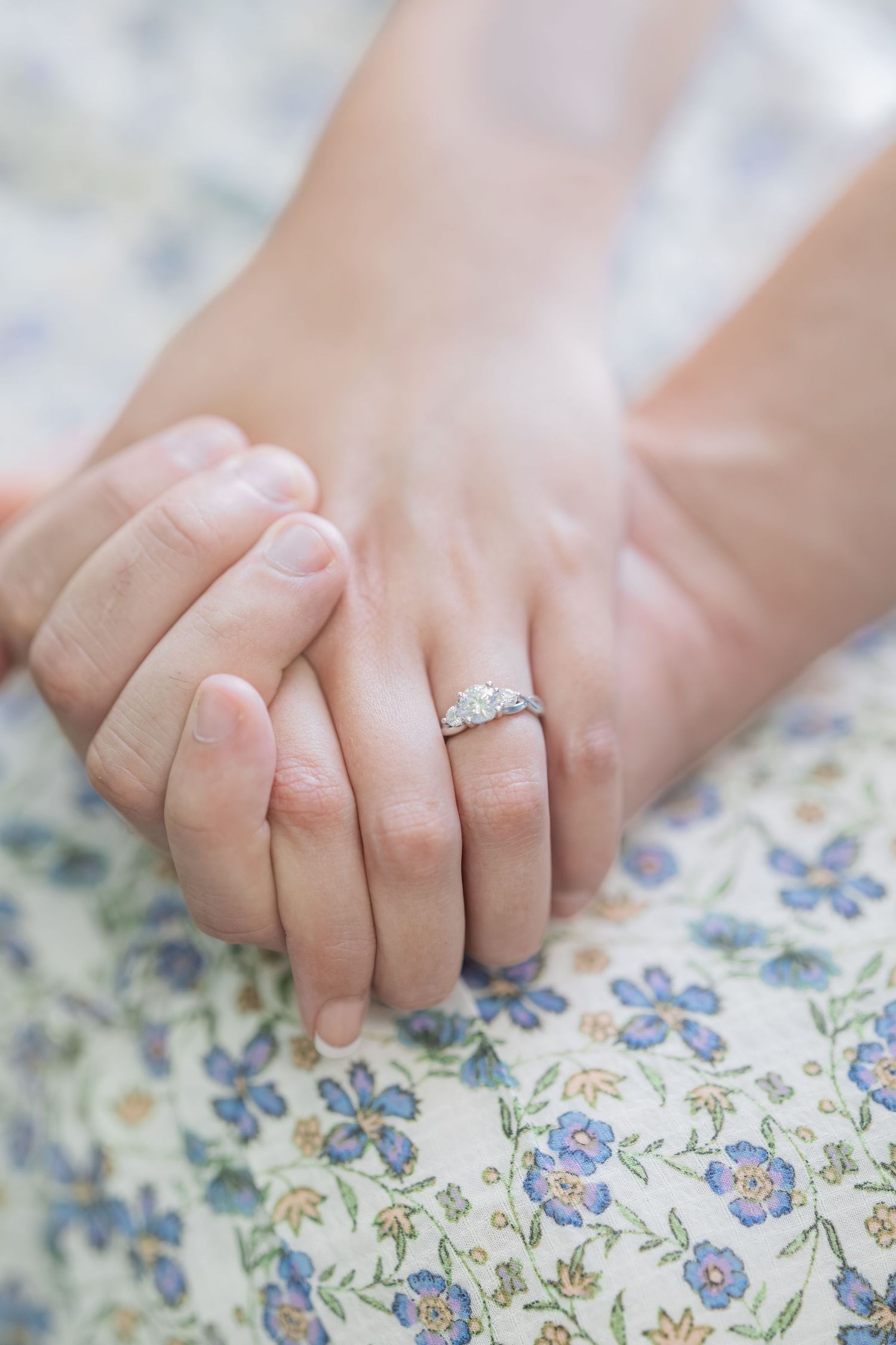 Close-up of couple holding hands with a diamond engagement ring on a floral fabric background.