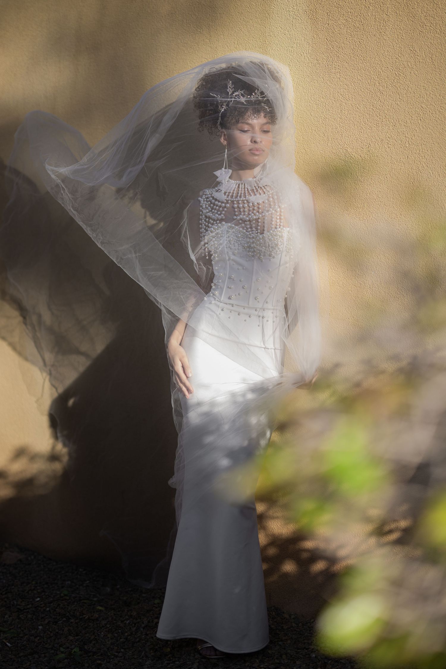 Bride in white lace gown and flowing veil with crystal tiara poses gracefully against a sunlit wall.