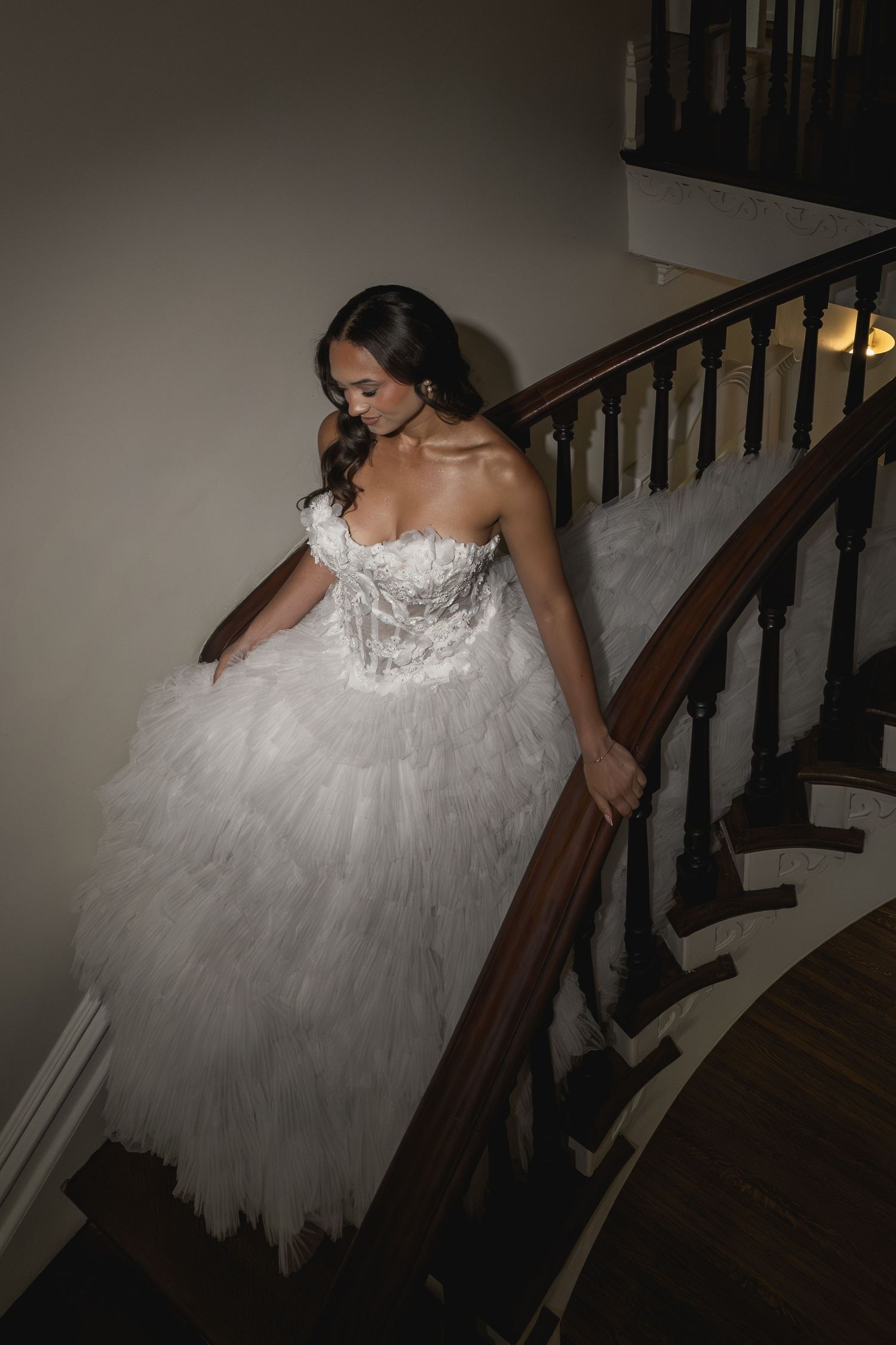 Bride in a strapless white tulle ball gown descending an elegant curved staircase with wooden railing.