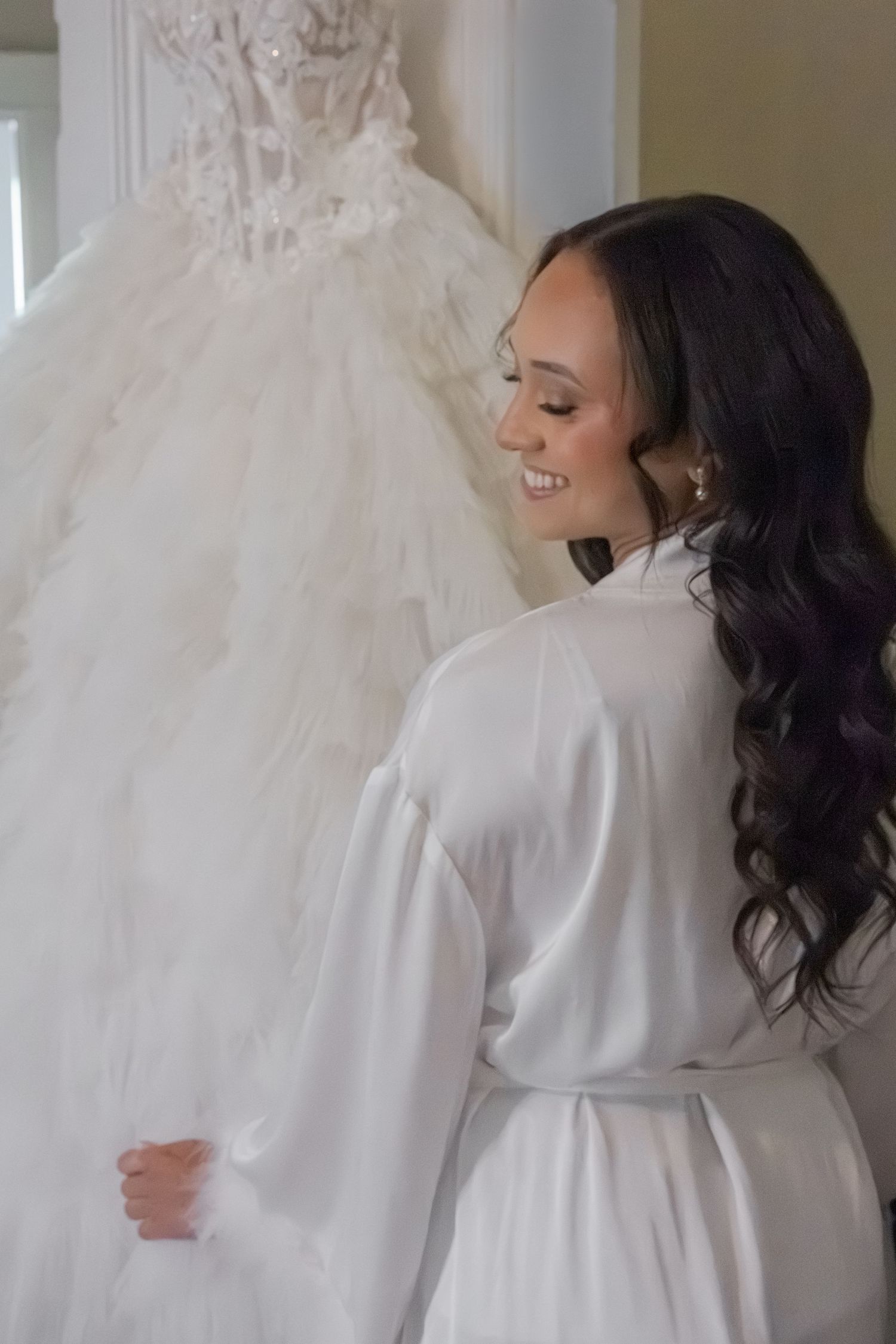 A smiling bride in a white satin robe admires her elegant lace and tulle ball gown wedding dress hanging on display.